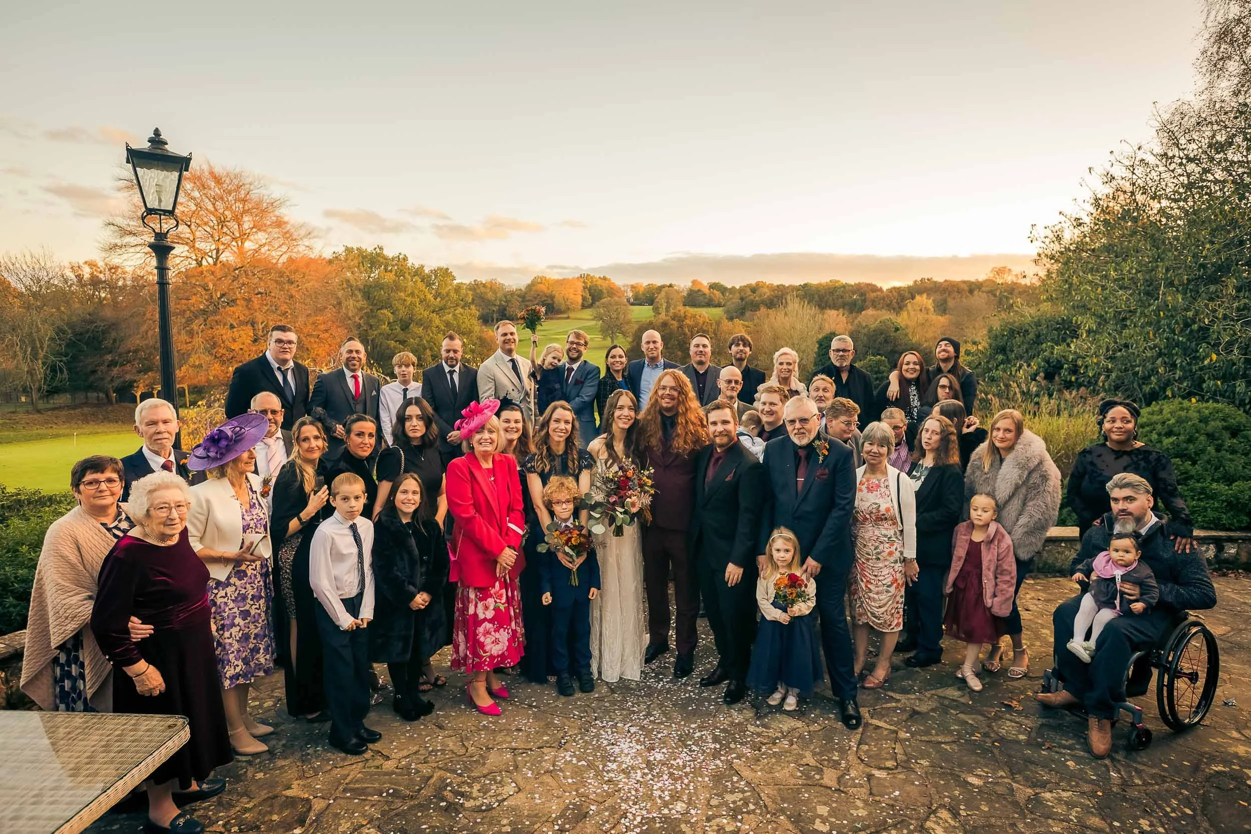 Large group of people gathered outdoors during sunset, celebrating a wedding. The bride and groom are at the center, surrounded by family and friends of different ages. The setting features a scenic backdrop of trees and a lawn.