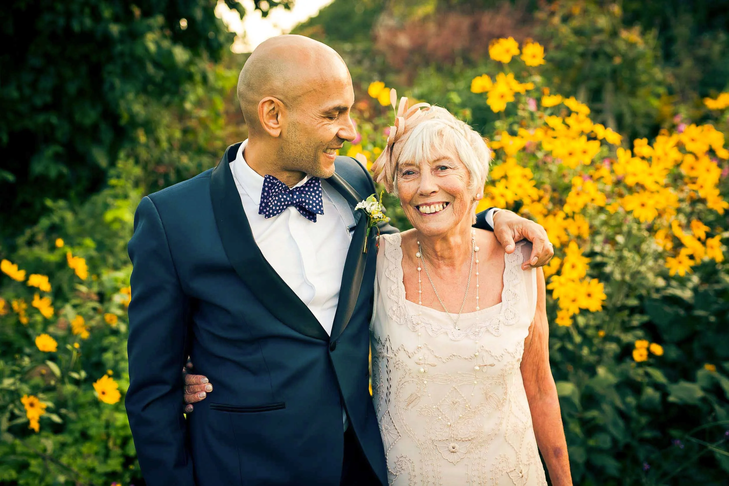 A man in a blue tuxedo with a polka dot bow tie hugging an elderly woman in a white lace dress with jewelry, both smiling, standing in front of yellow flowers in a garden.