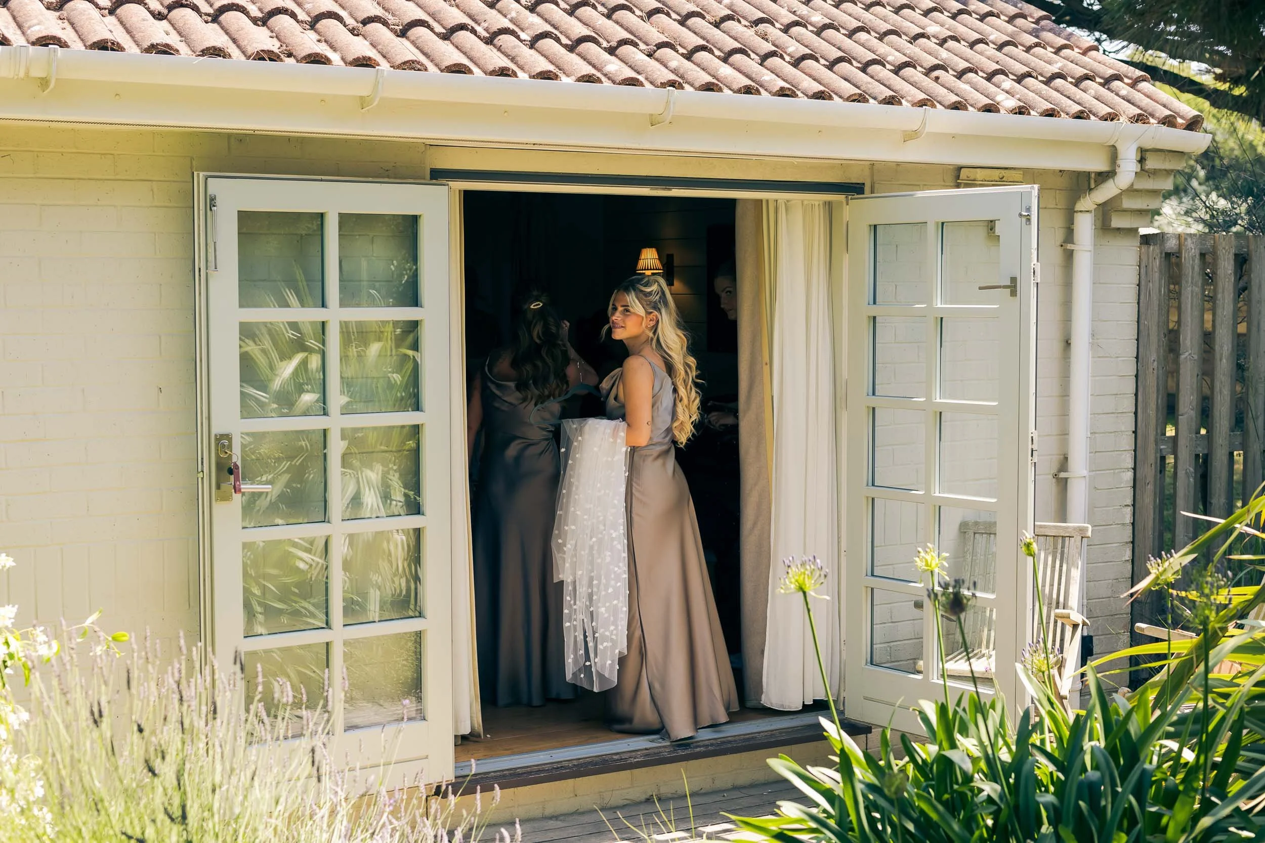 Two women in elegant dresses standing at an open French door leading outside, with sunlight and greenery visible.