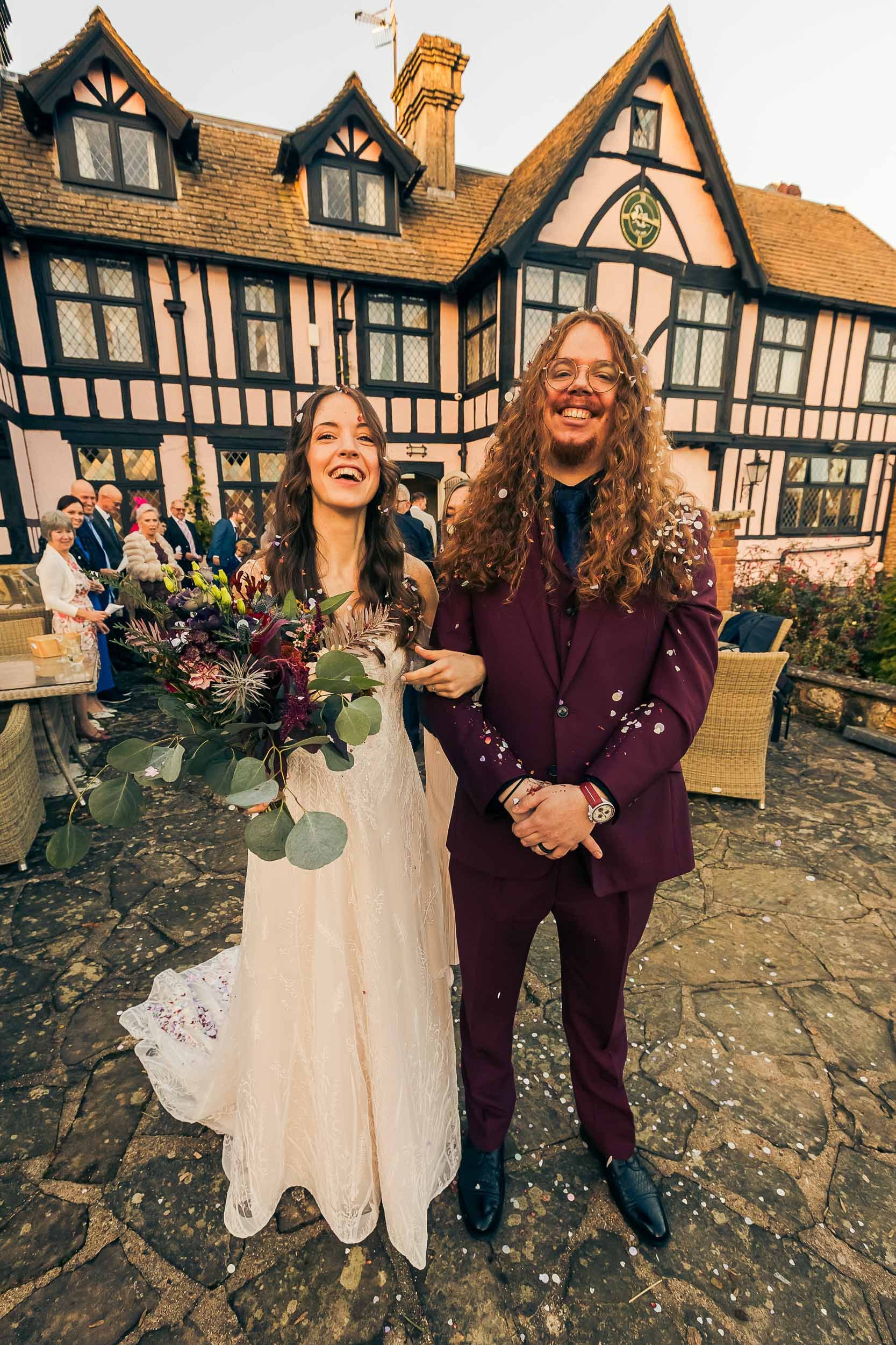 A bride and groom at their wedding celebration outdoors in front of a Tudor-style house, with guests in the background, confetti falling around them, and joyous expressions.