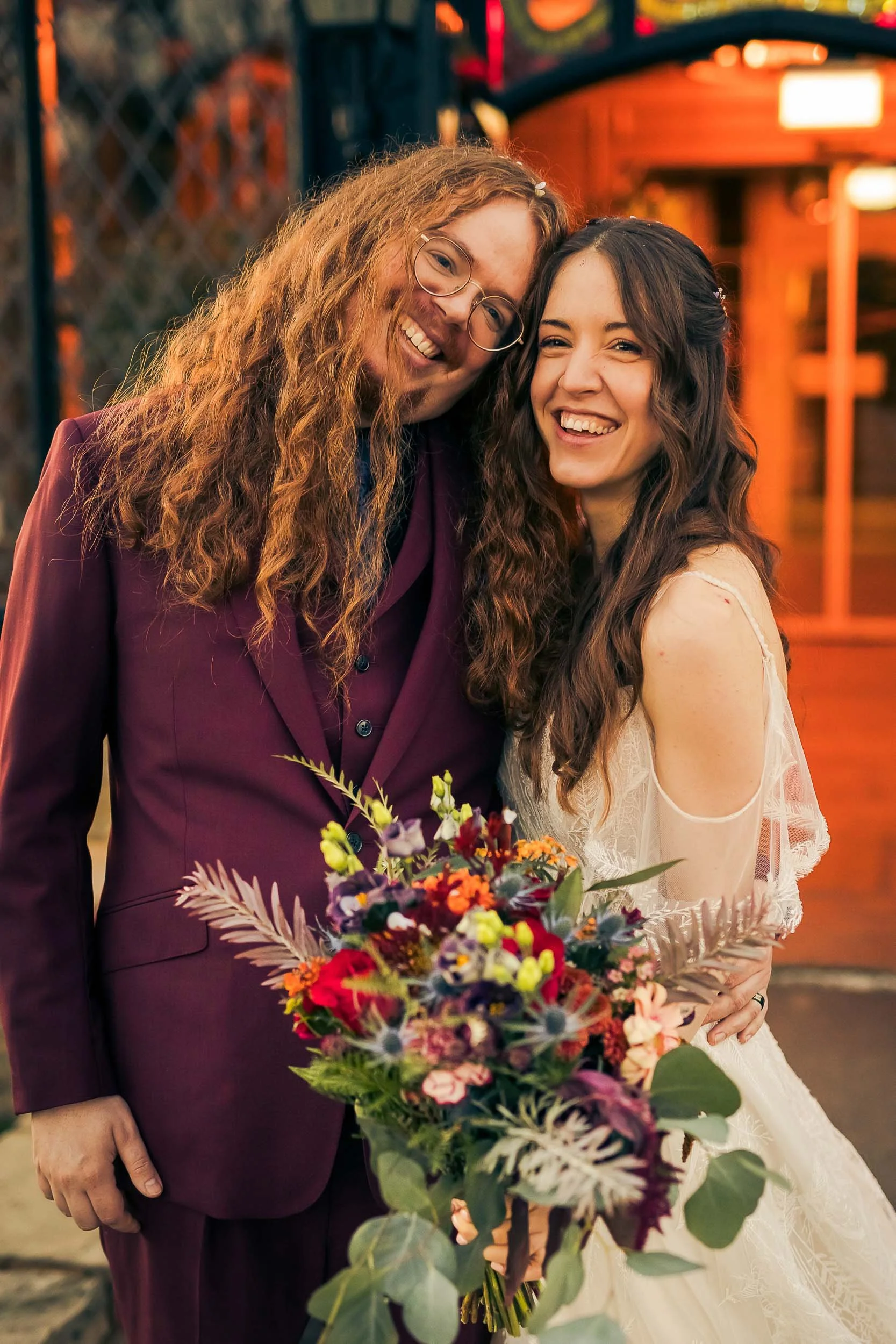 Two women smiling and embracing at a wedding, one holding a colorful bouquet of flowers, in an outdoor setting with warm lighting and decorative background.