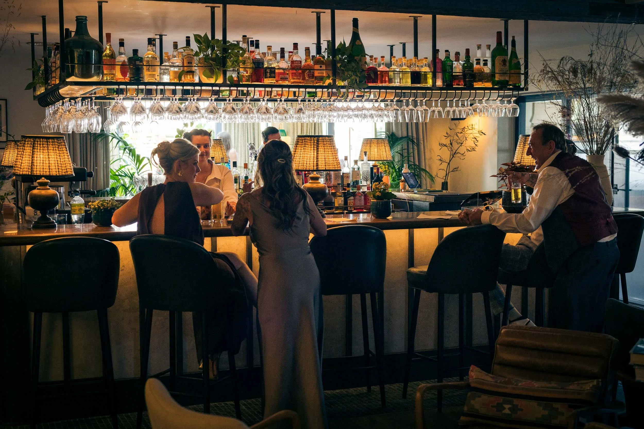 People sitting at a bar in a cozy, dimly-lit restaurant or bar with bottles of alcohol and glasses hanging above the counter.