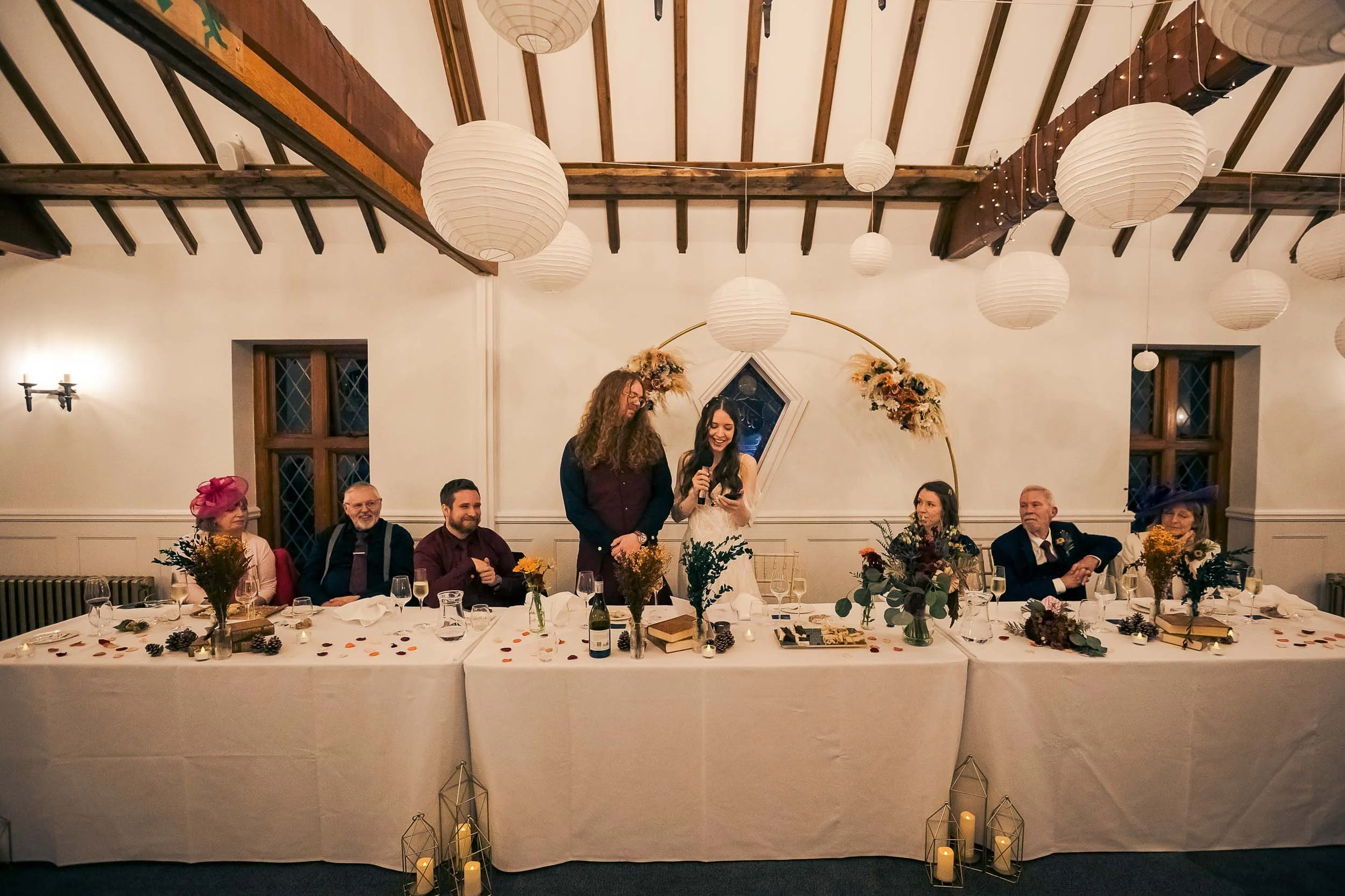 A bride giving a speech at a wedding reception table with seven seated guests, floral centerpieces, and decorative lighting in a rustic decorated hall.