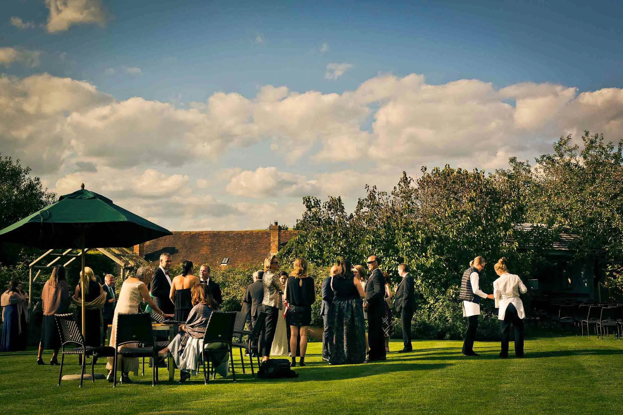 People gathered outdoors in a garden for a social event, with some seated at tables and others standing and chatting, under a large green umbrella, with trees and a house with a tiled roof in the background.
