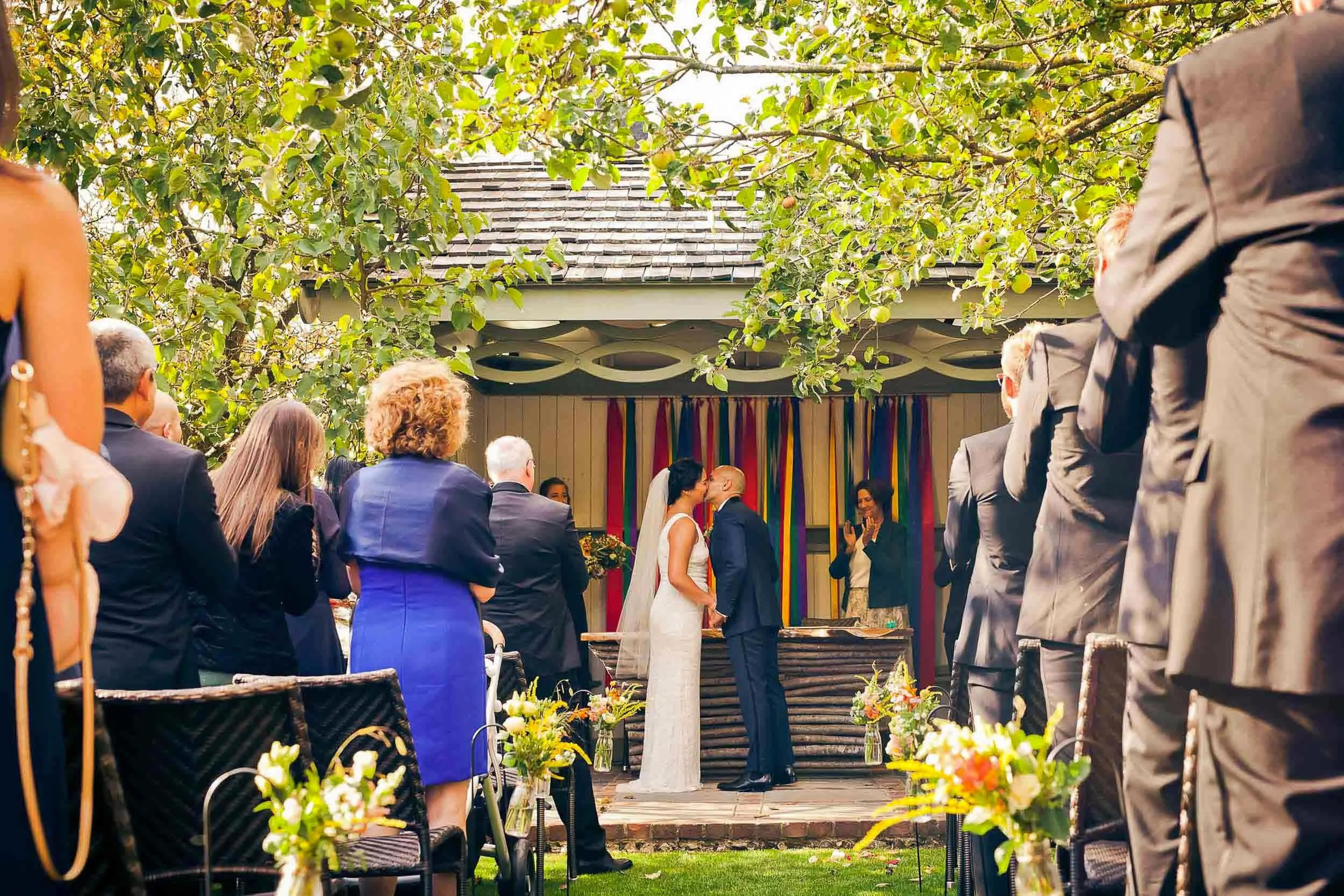 A outdoor wedding ceremony with the bride and groom sharing a kiss at the altar, surrounded by guests standing and seated, with a backdrop of colorful vertical ribbons hanging on the building behind.