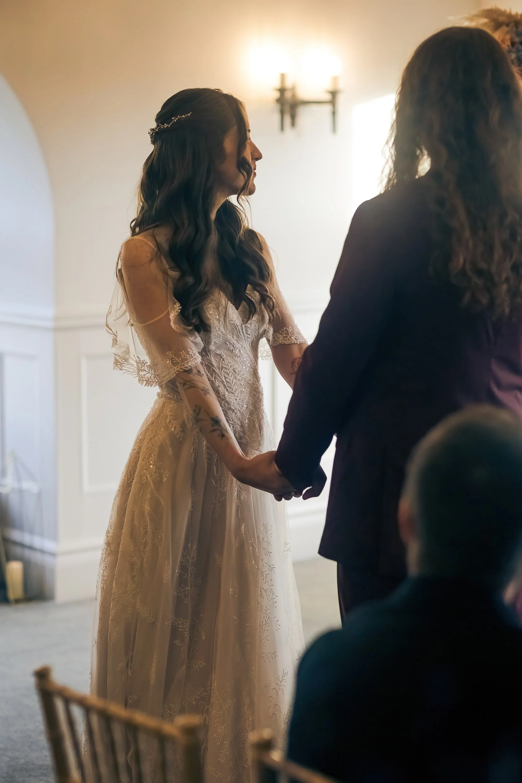 A bride in a lace wedding dress holding hands with another woman during a wedding ceremony.