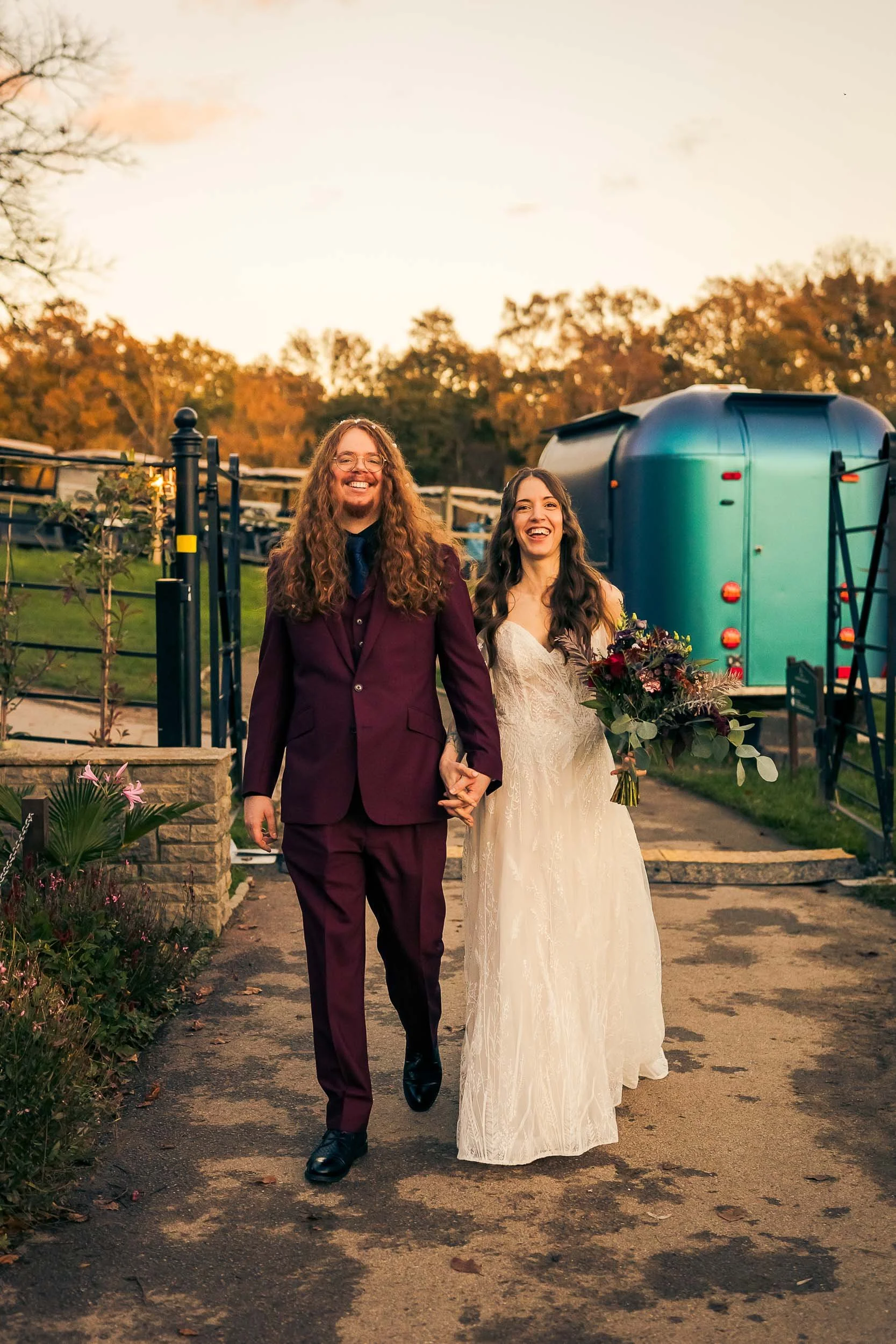 A newlywed couple walking hand-in-hand outdoors at sunset, smiling and happy, with the bride holding a bouquet of flowers.