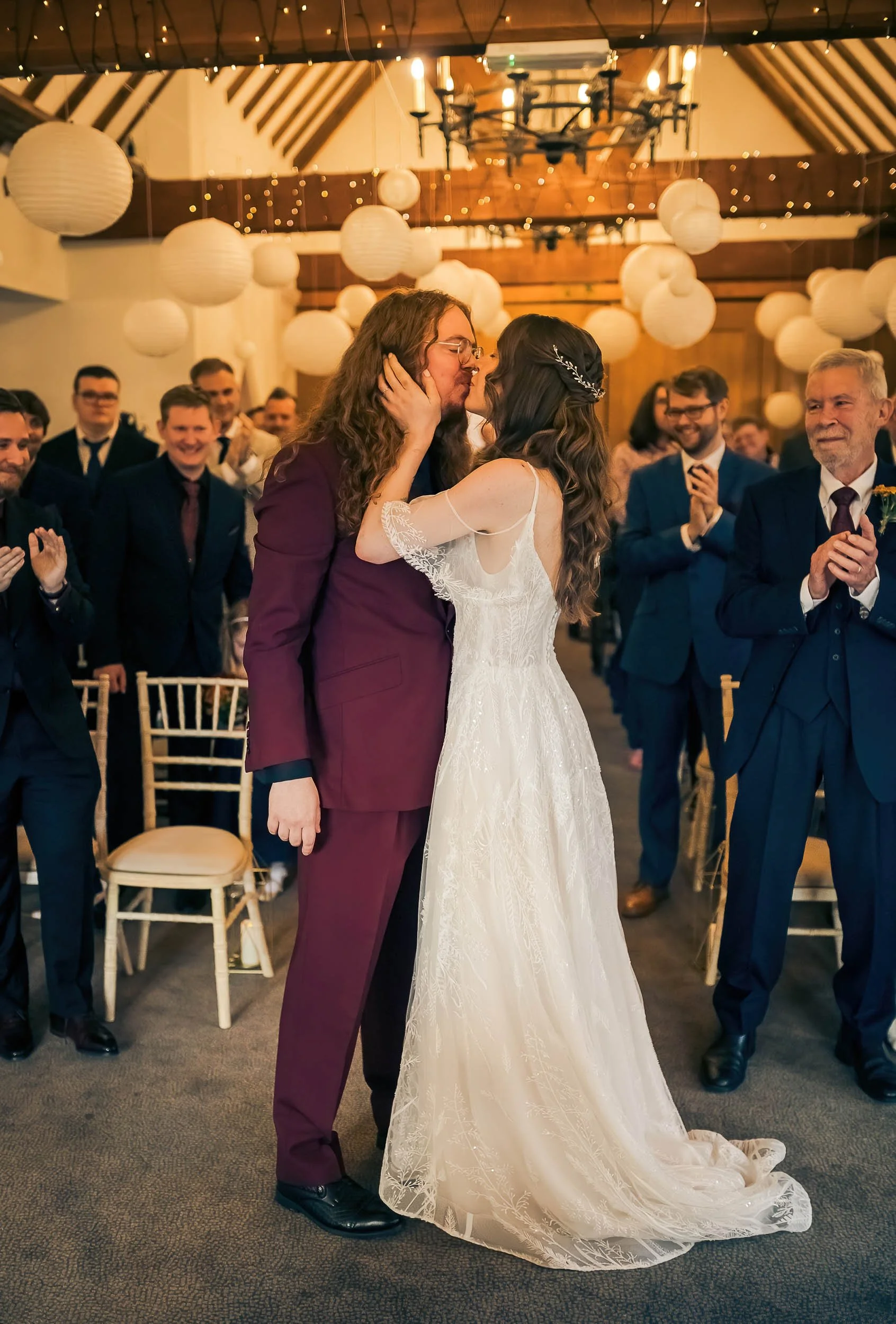 A couple shares a kiss at their wedding ceremony while surrounded by family and friends in a decorated venue with hanging lanterns and fairy lights.