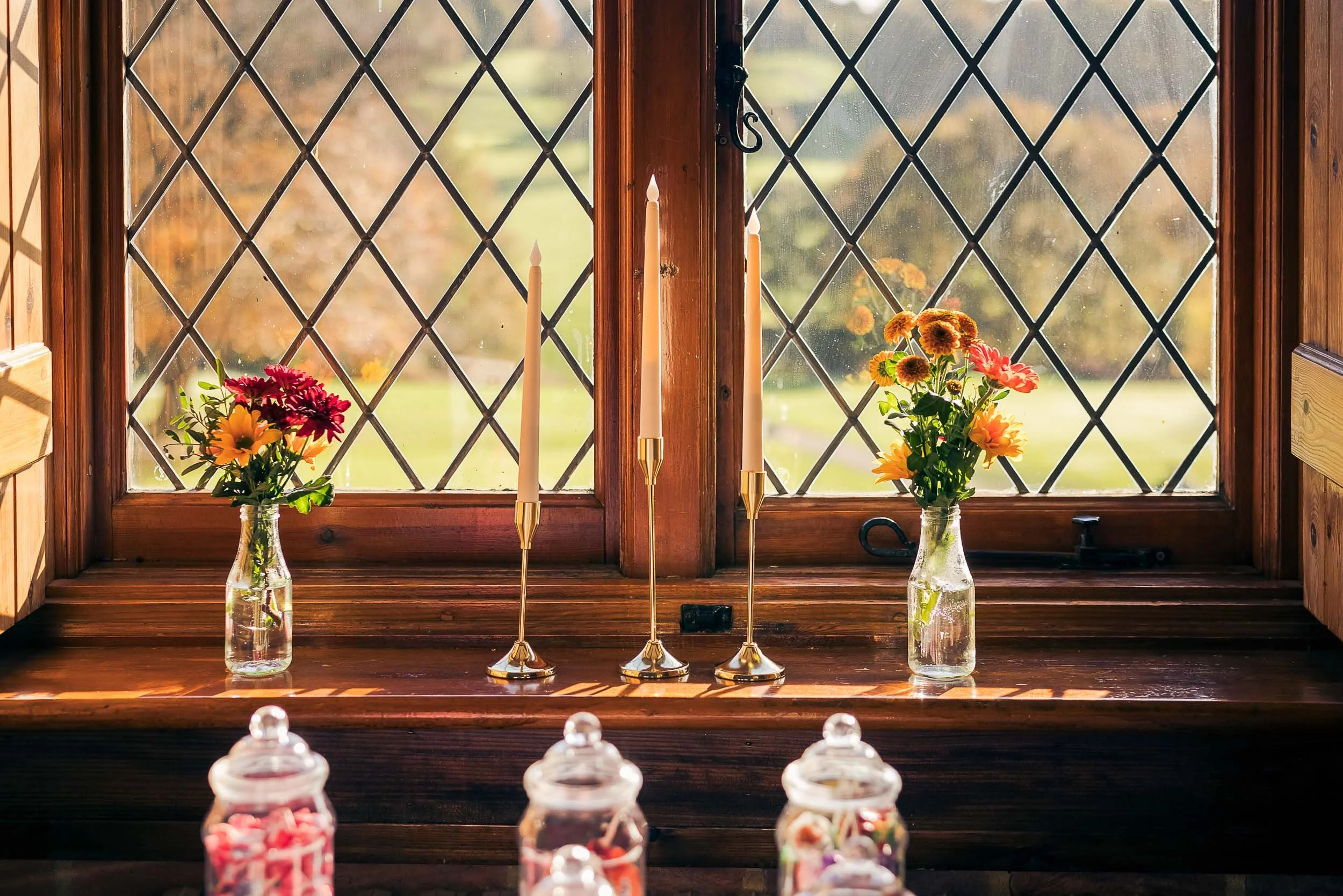 Sunlight streaming through a diamond-patterned window onto a wooden windowsill with two glass vases containing colorful flowers and four gold candlesticks, in a cozy rustic setting.