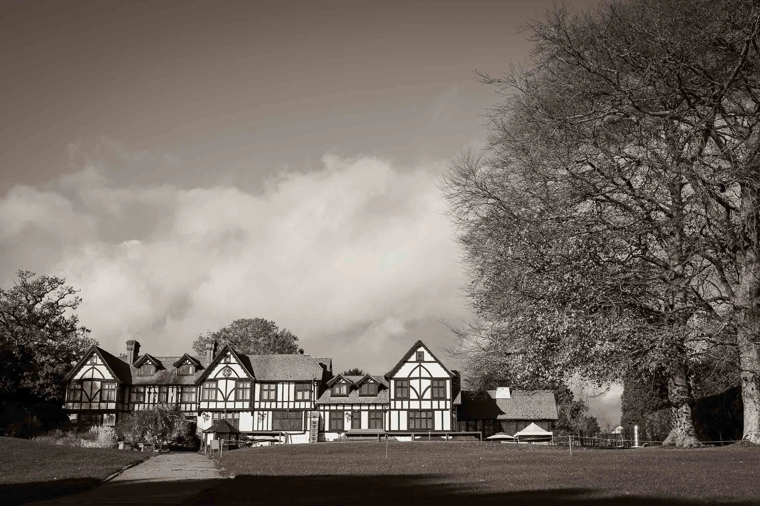 A large multi-story house with Tudor-style architecture, surrounded by a grassy yard with a paved pathway, large trees with bare branches, and a cloudy sky in the background.
