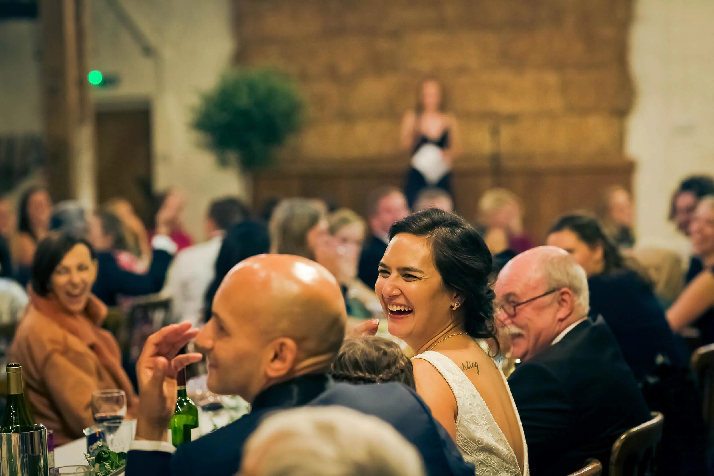People laughing and enjoying a dinner event, with a woman in the center wearing a white dress, surrounded by guests in formal attire, in a warmly lit room with wooden walls.