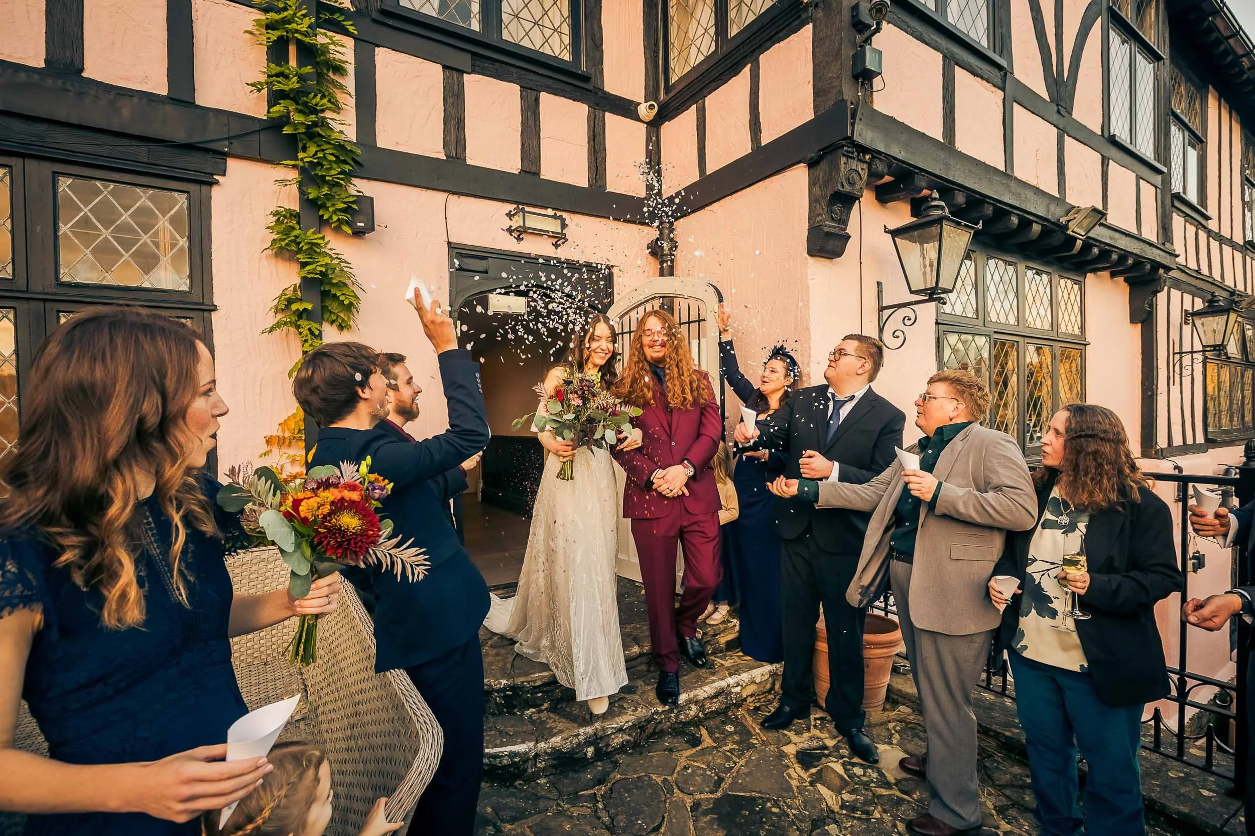 A wedding celebration outside a quaint, half-timbered building with guests throwing confetti as the bride, wearing a white dress, and the groom, in a maroon suit, smile at each other. Guests hold bouquets and glasses of wine.