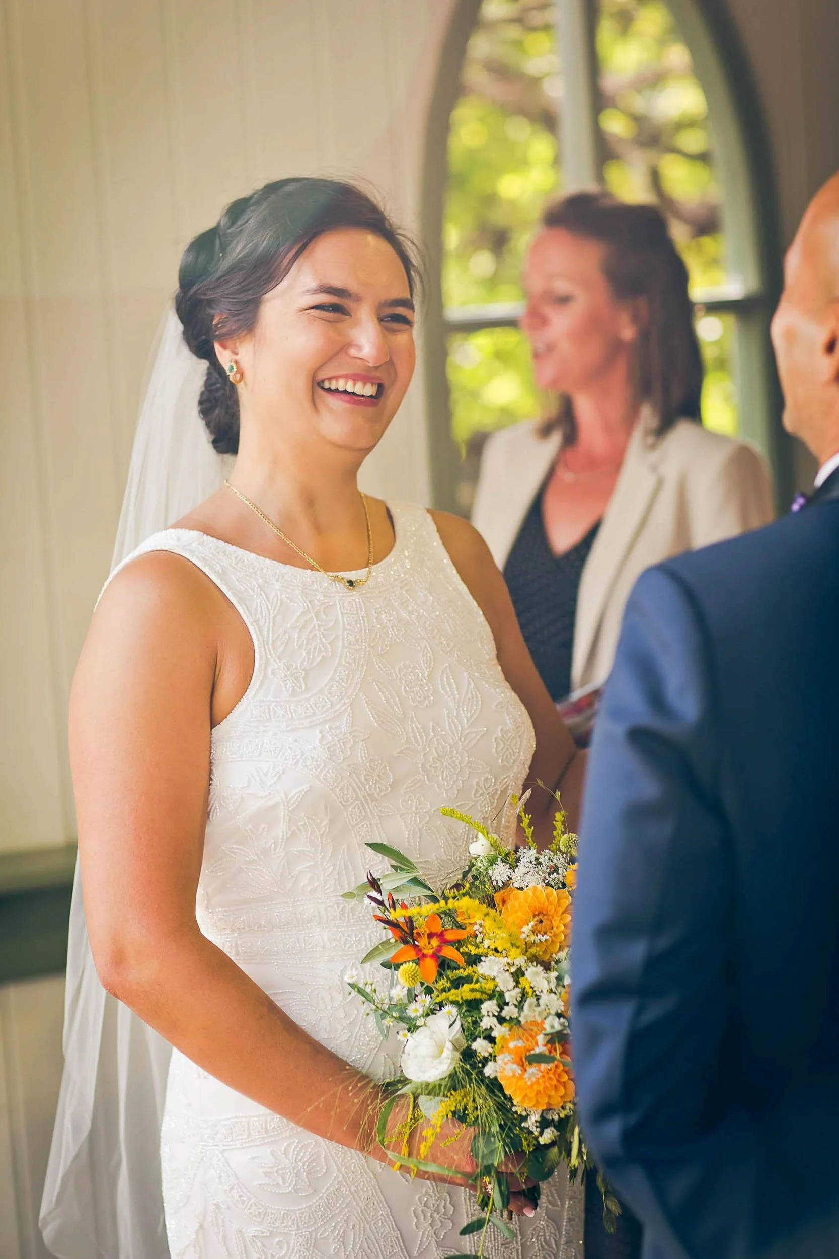 A smiling bride in a white dress holding a bouquet of yellow and orange flowers during her wedding ceremony.