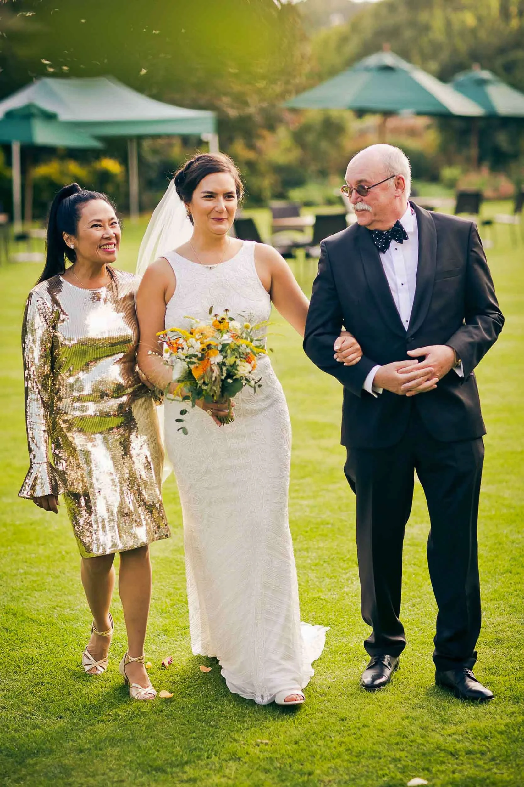 A bride in a white lace wedding gown holding a bouquet, walking with a man in a black suit and a woman in a shiny gold dress, outdoors on a grassy area with umbrellas in the background.
