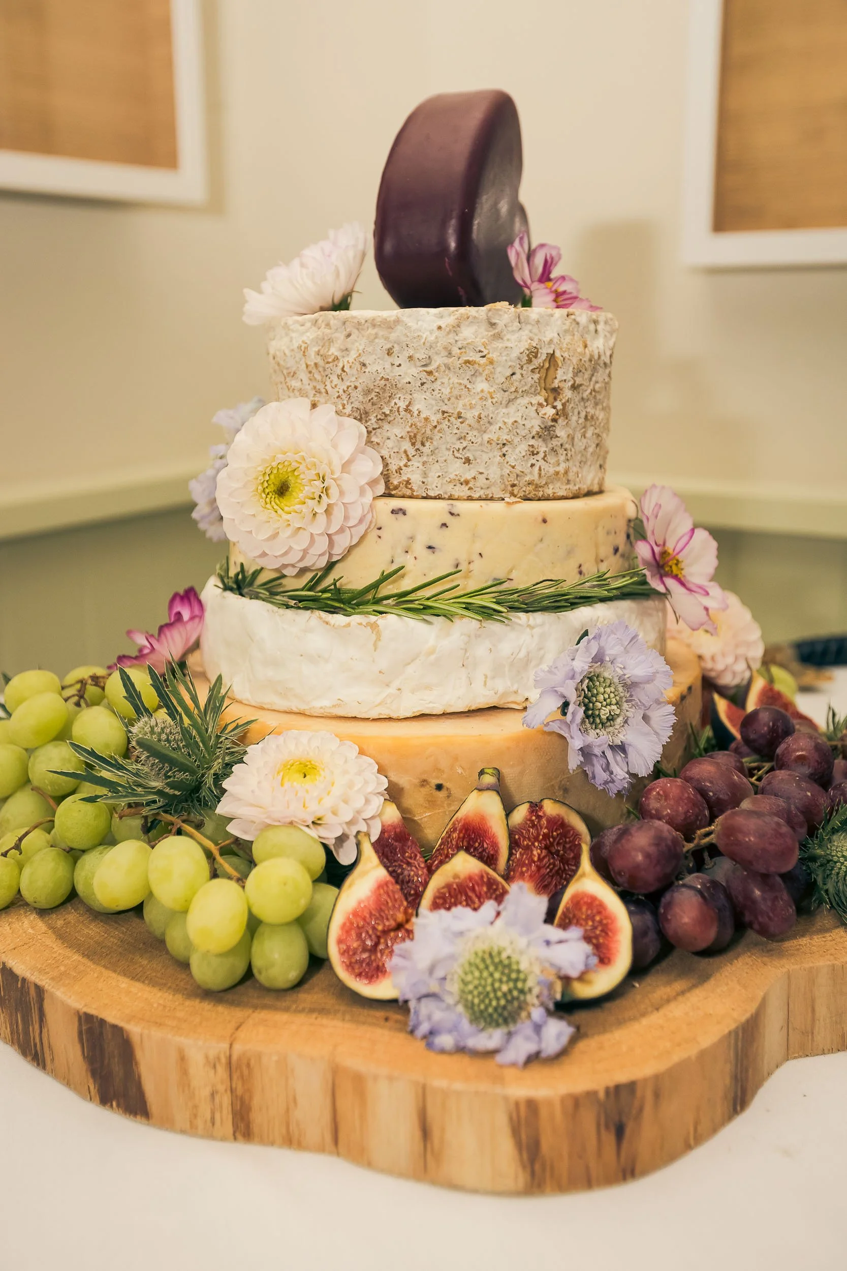 A multi-tier cheese cheese cake decorated with flowers and surrounded by grapes and figs on a wooden board.