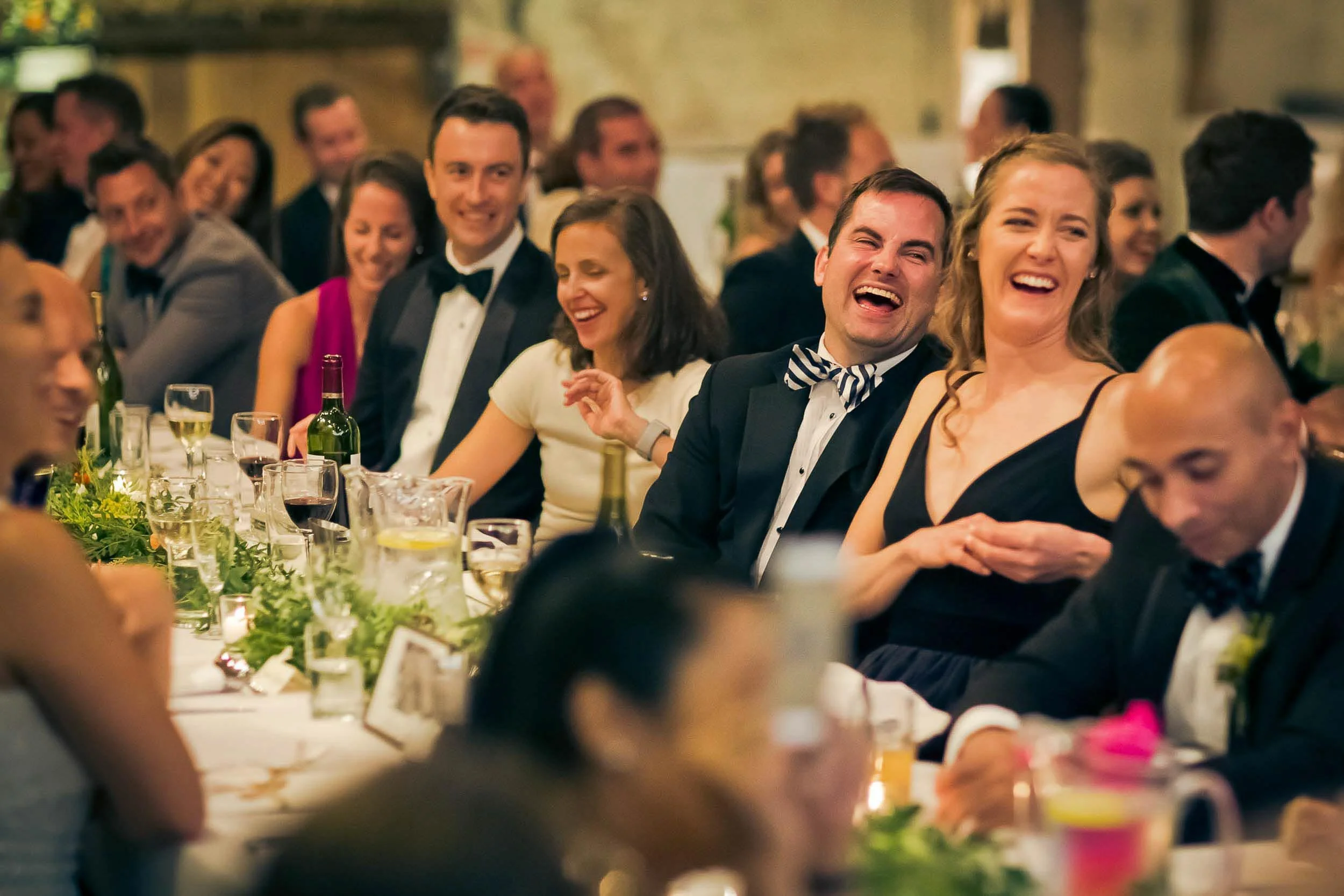 People in formal attire laughing and enjoying at a dinner event with drinks and floral decorations on the table.