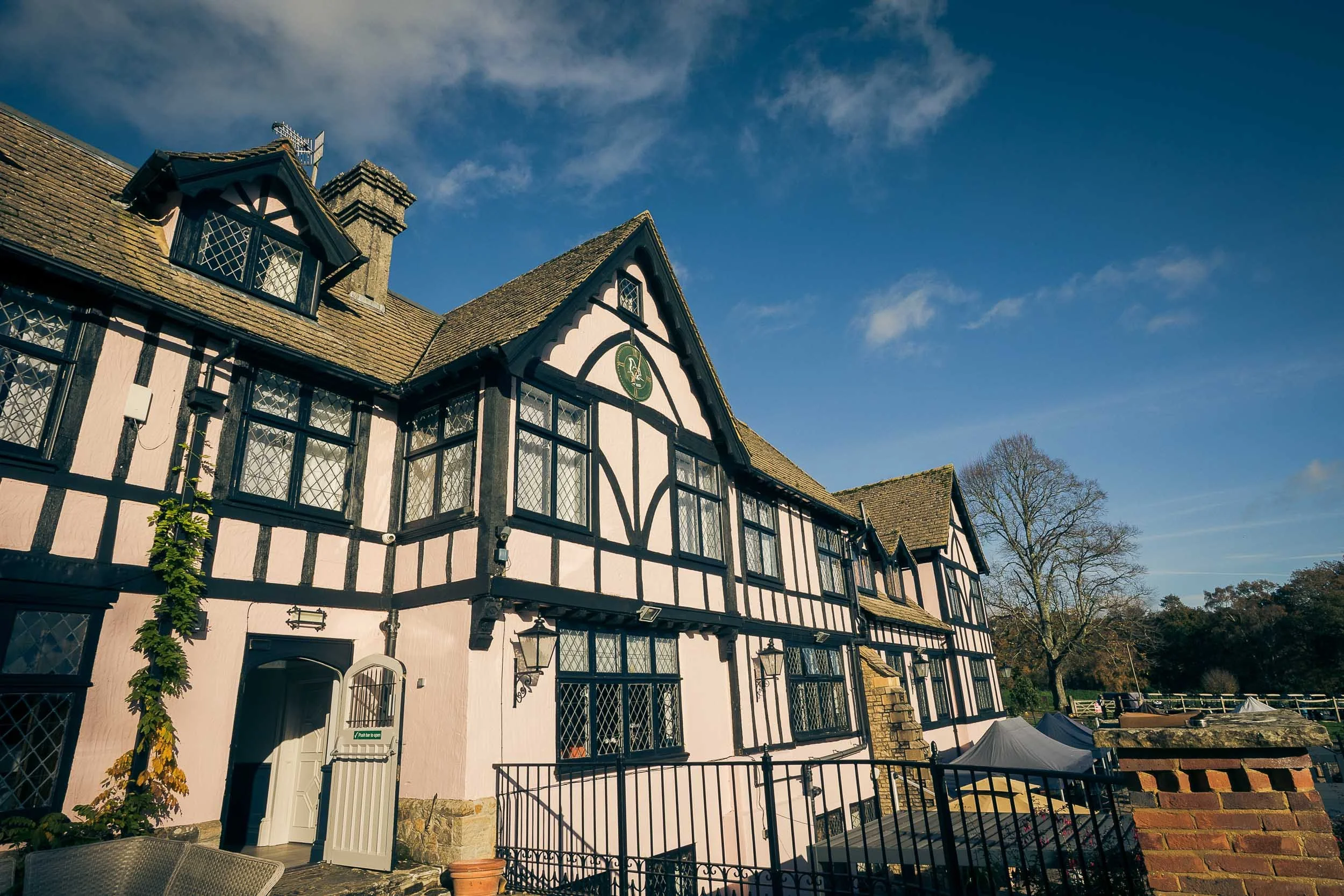 A large, historic Tudor-style building with black timber framing and white infill, multiple windows with diamond-pane glass, and a steeply pitched roof under a blue sky with clouds.
