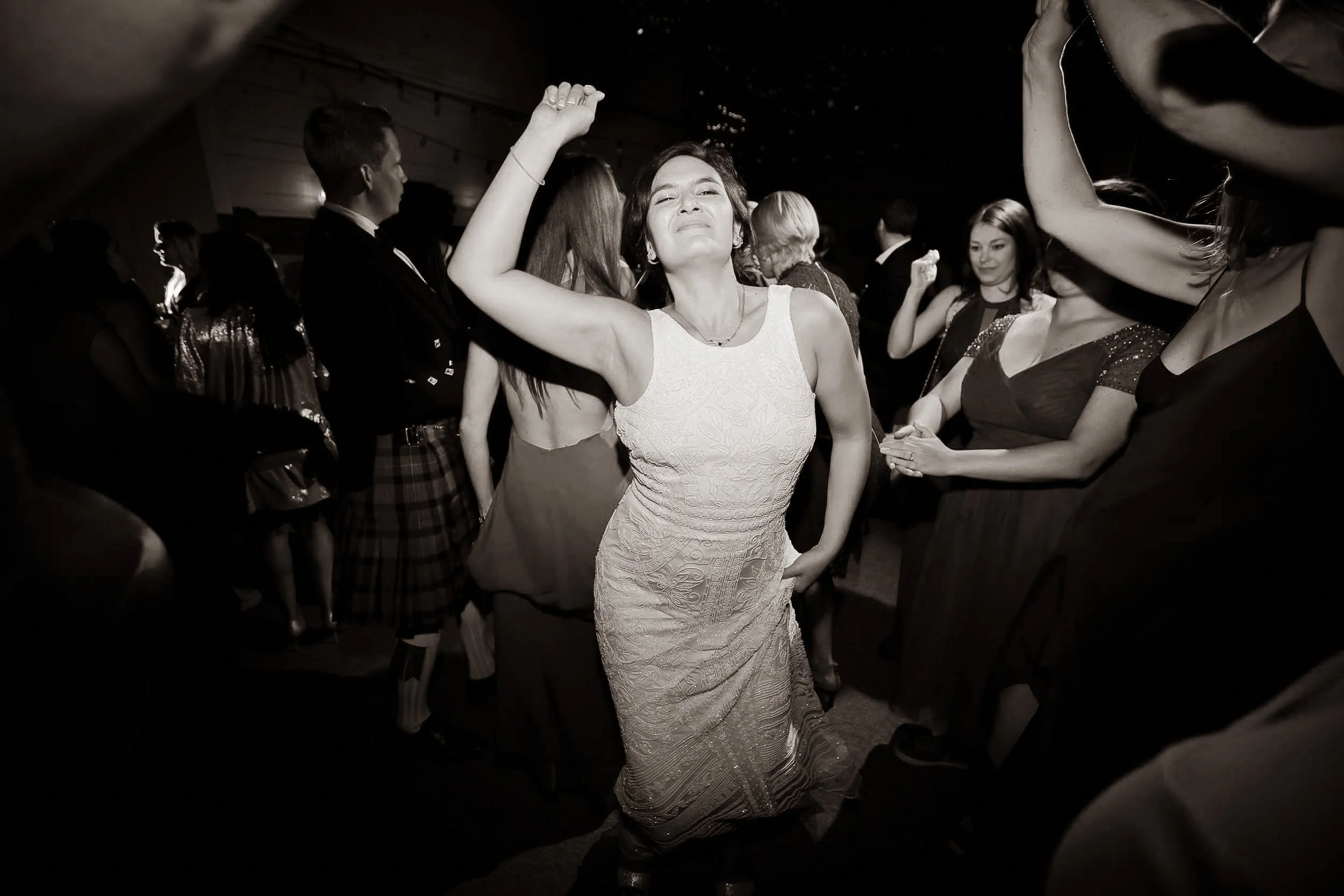 A woman in a sleeveless, patterned dress dancing at a party, surrounded by other people, some dancing and others watching, in a dark indoor setting.