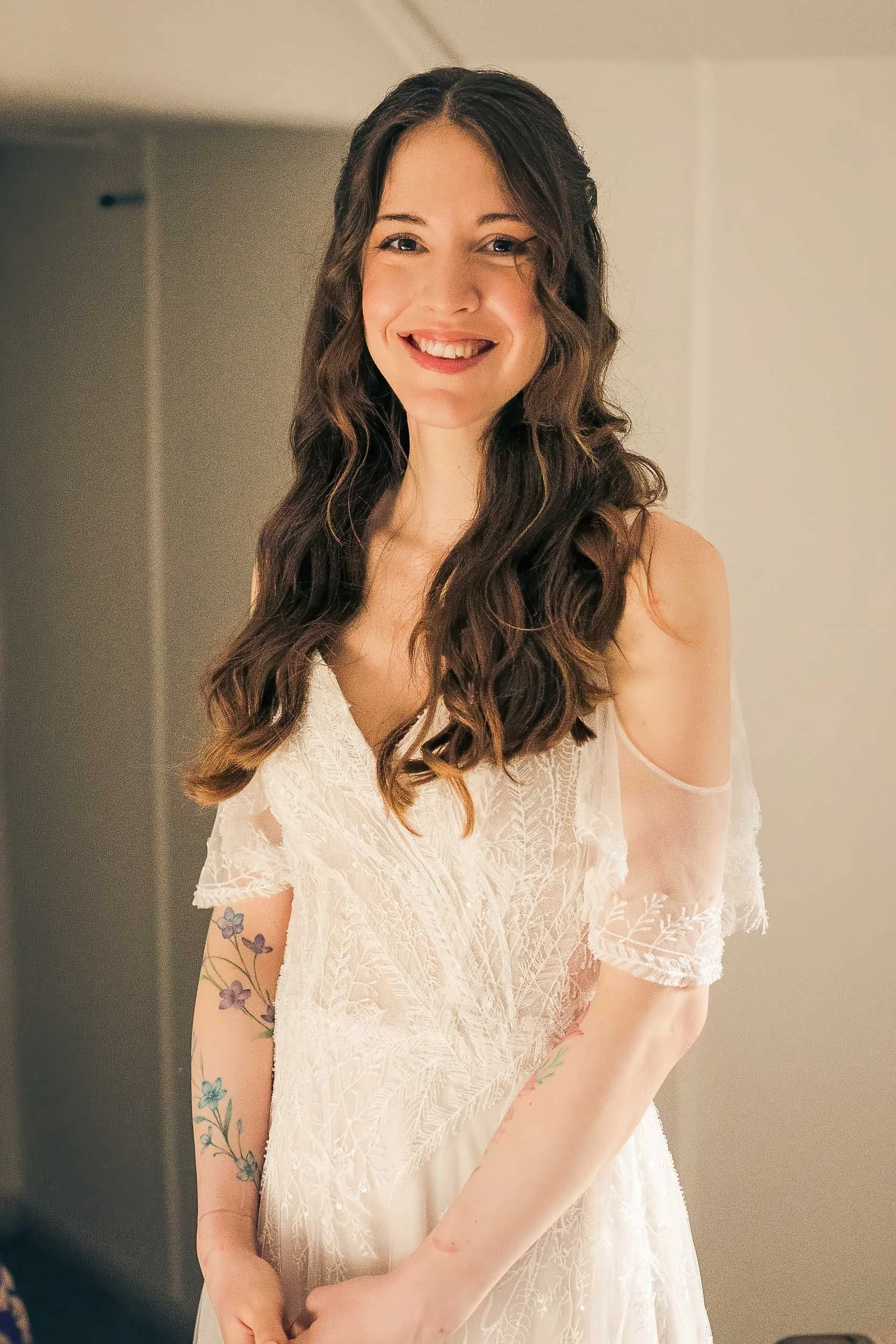 A young woman with long, wavy brown hair and a tattoo on her arm, wearing a white lace dress, smiling at the camera.