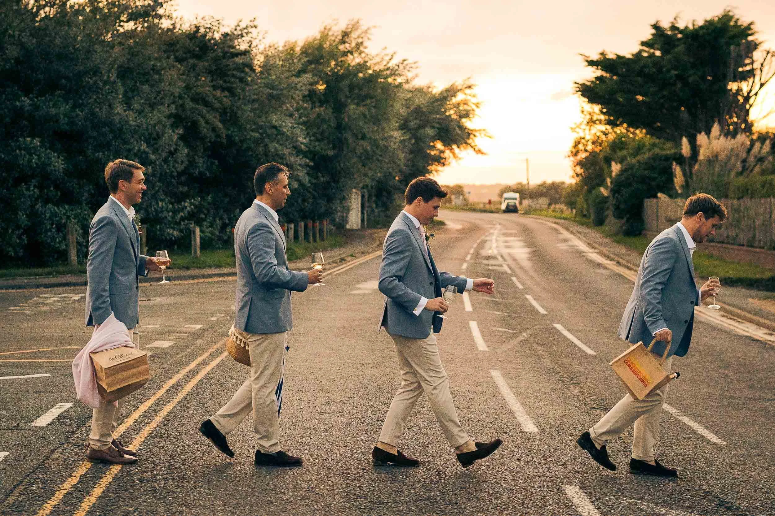 A humorous recreation of The Beatles' Abbey Road album cover, with five young men in suits walking across a road at sunset, each holding a drink and carrying shopping bags.