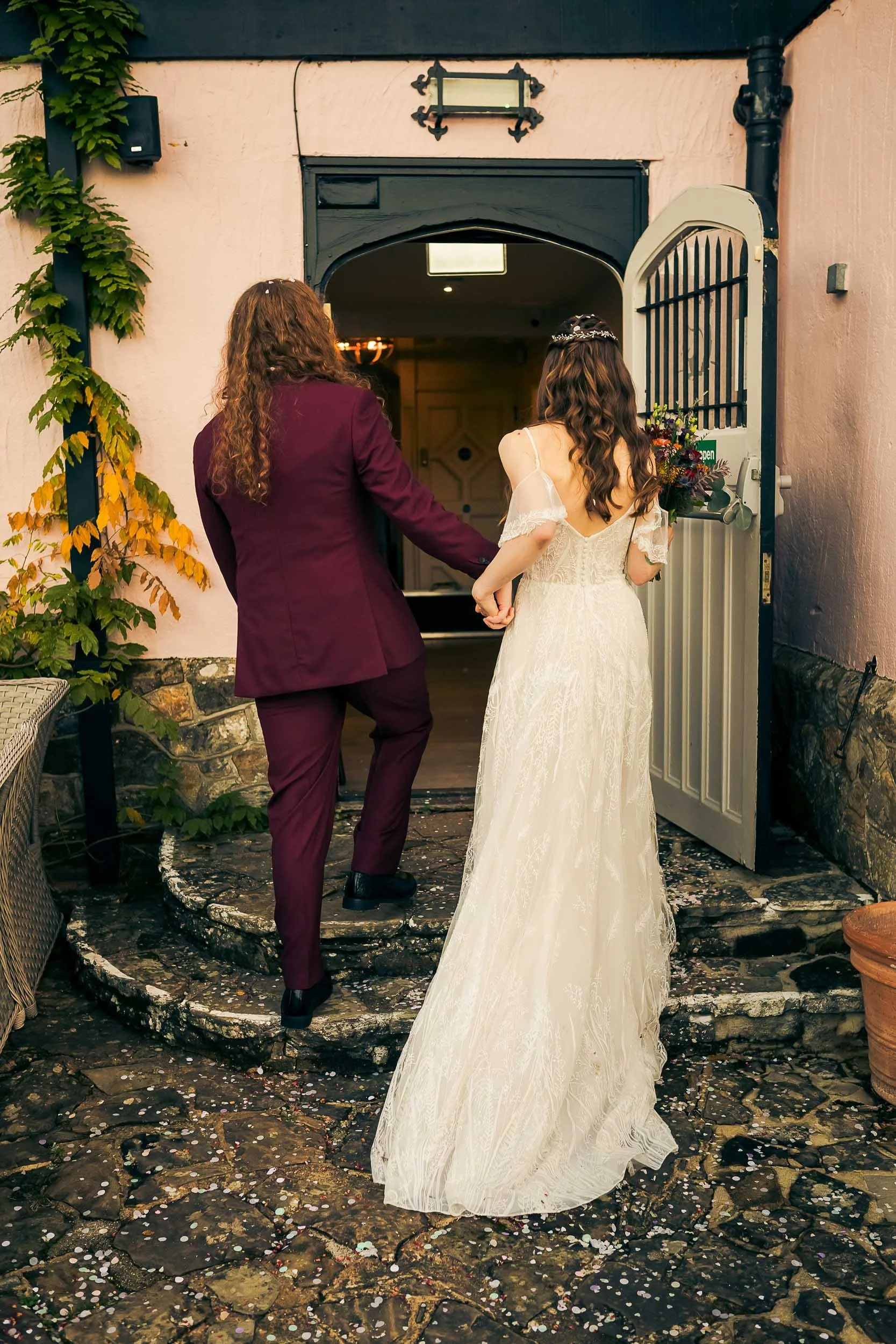 A bride in a white wedding dress and a person in a maroon suit walking hand-in-hand into a building, likely for a wedding ceremony or celebration.