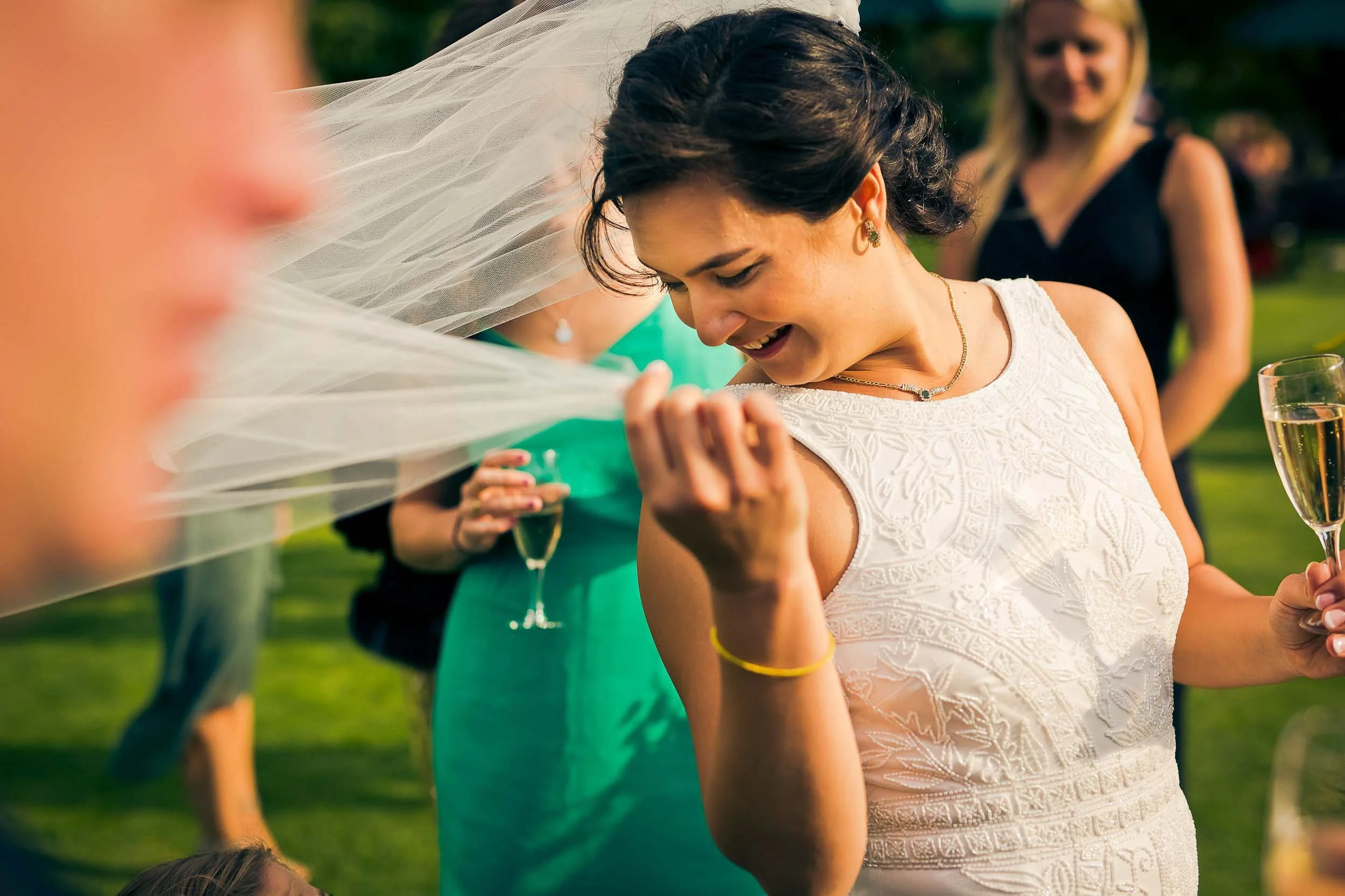 A woman in a white embroidered dress holding a champagne glass at an outdoor wedding reception, smiling and looking at her shoulder veil, with other guests in the background.