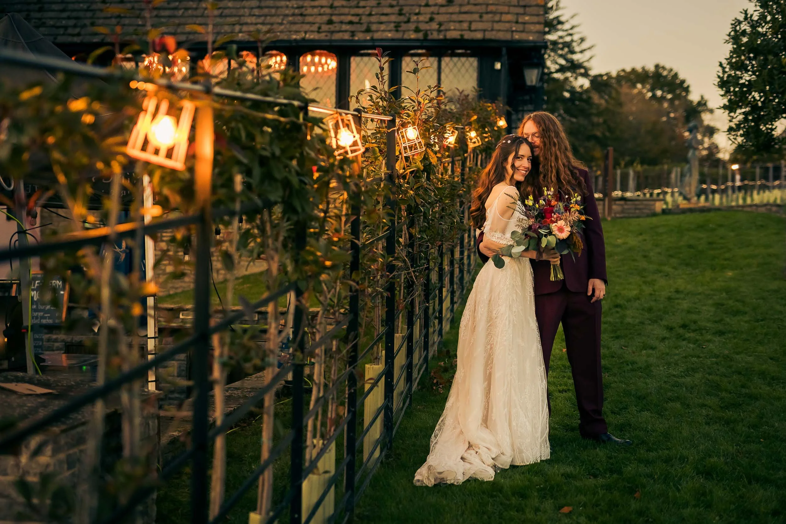 A bride and groom smiling and embracing outdoors during sunset, with the bride holding a bouquet of flowers, standing near a fence covered with string lights.