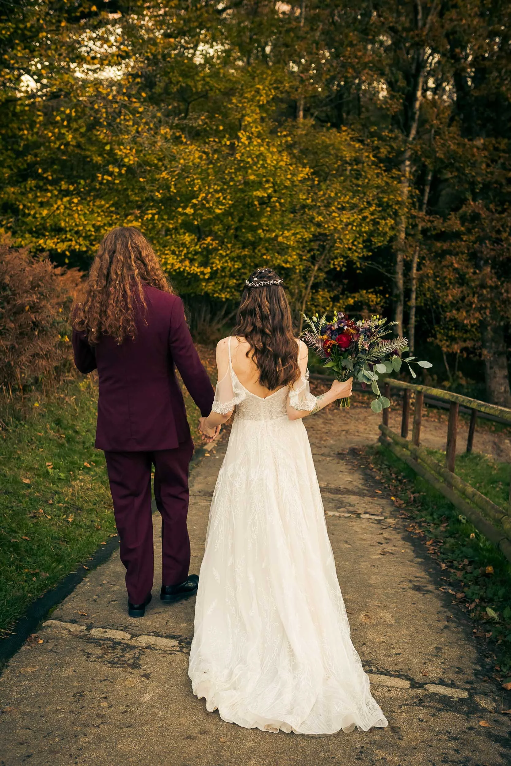A couple, dressed in wedding attire, walking hand in hand on a path through a park during autumn. The bride is holding a bouquet of flowers.