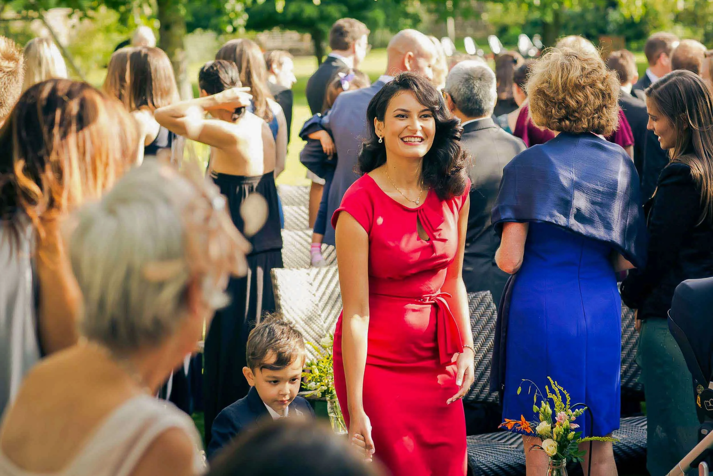A cheerful woman in a red dress standing in a crowd at an outdoor event, smiling at the camera, with other people in the background.