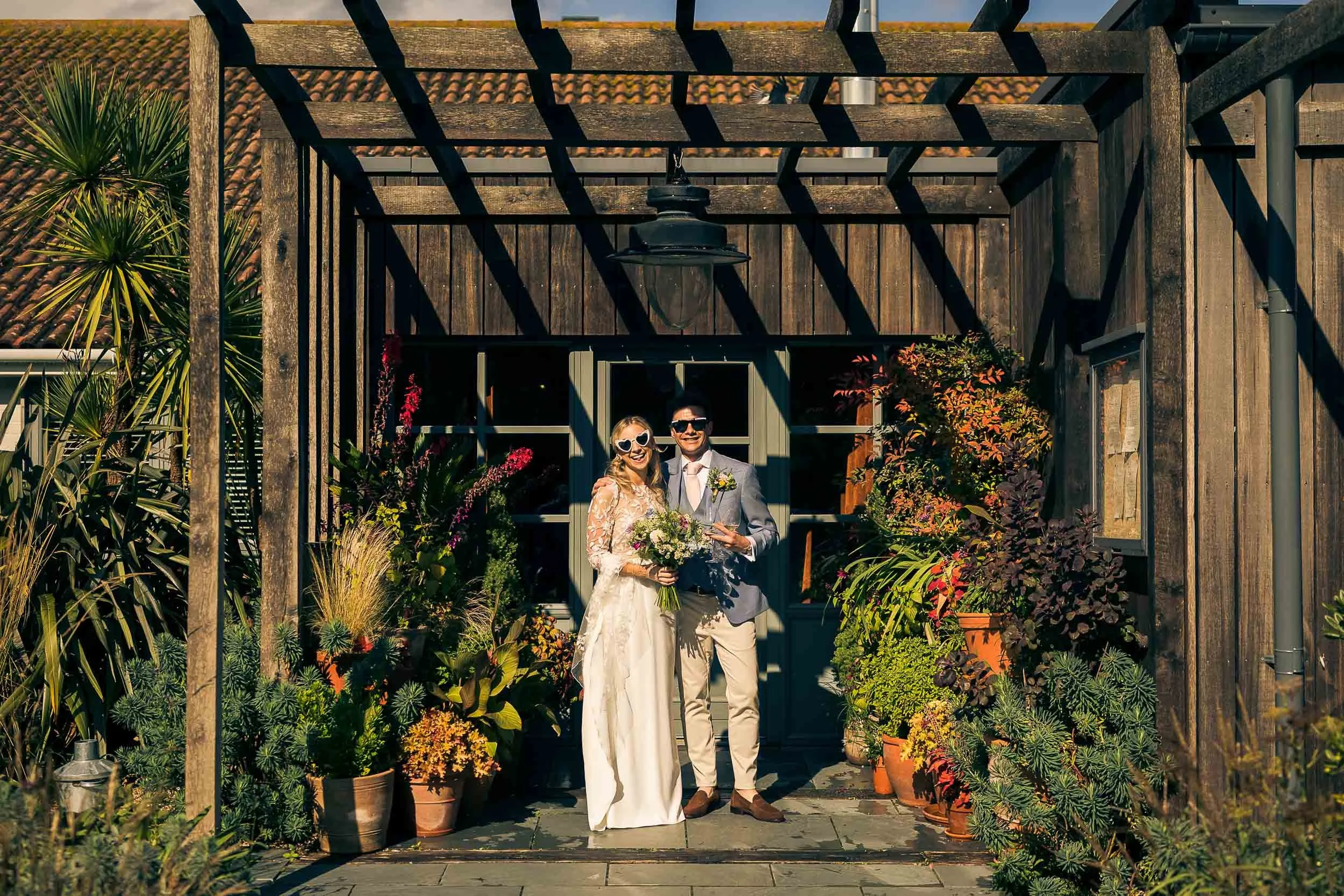 A newlywed couple stands in front of a rustic wooden building, smiling. The bride holds a bouquet of flowers, and both are dressed in wedding attire. They are surrounded by potted plants and flowers, with sunlight casting shadows.