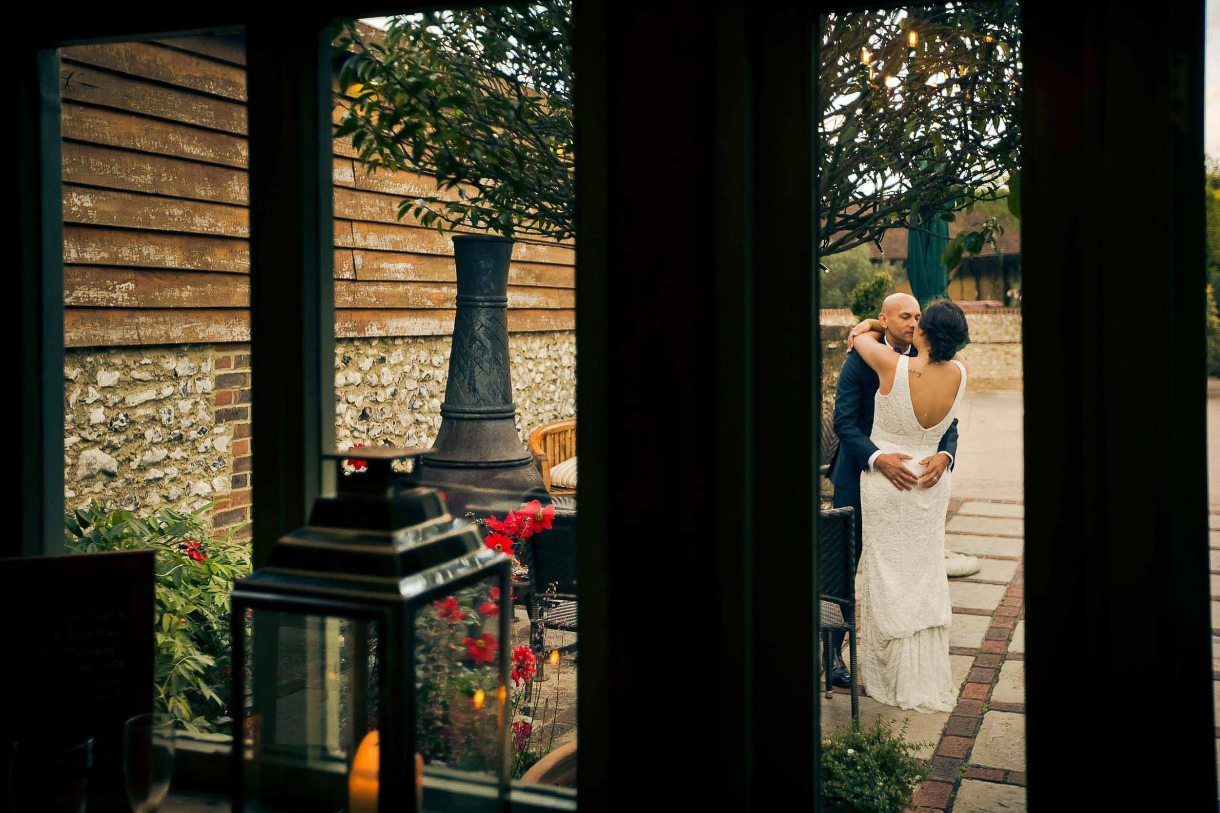A couple dancing outdoors, as viewed through a window, with the man wearing a dark suit and the woman in a white dress, surrounded by an outdoor garden with plants and a stone pathway.