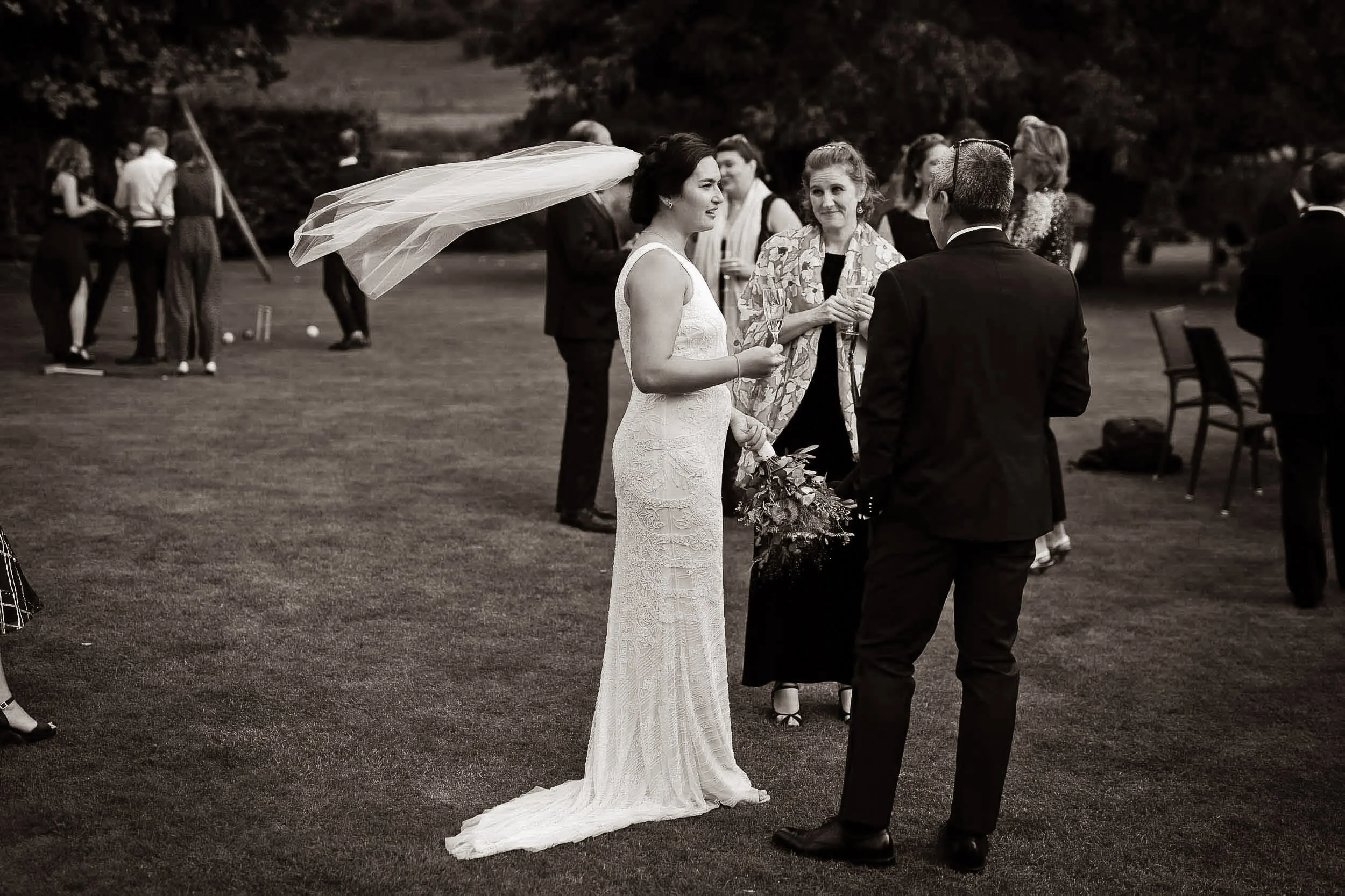 A bride in a lace wedding gown holding a bouquet, standing outside on grass, speaking with a groom in a black suit, with guests mingling in the background.