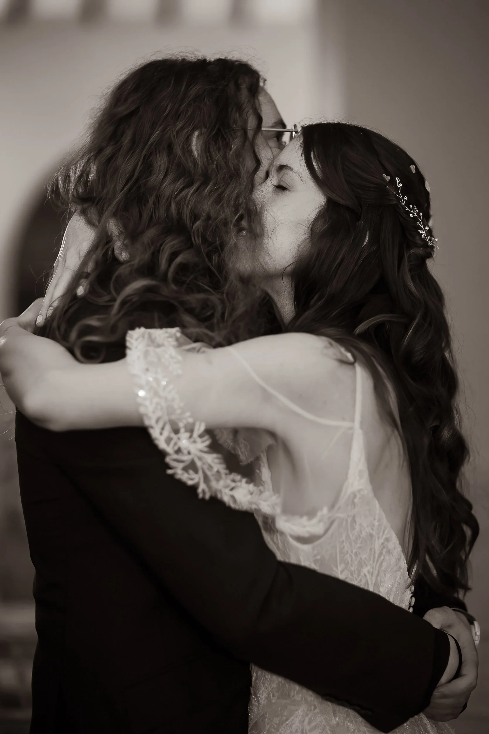 A black and white photo of two women embracing and kissing, one with curly hair and glasses, the other with long wavy hair and a decorative hairpiece, dressed in wedding attire.