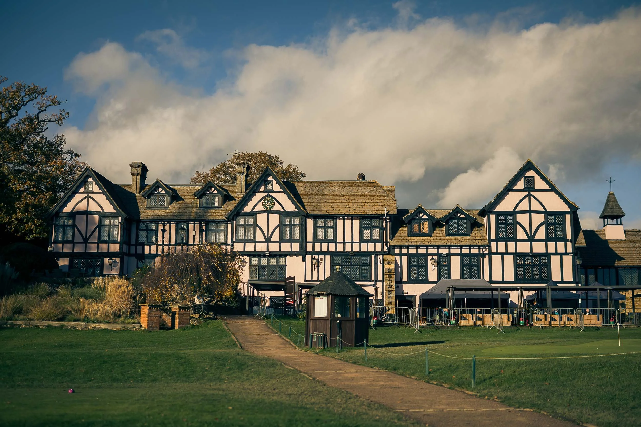 A large, traditional Tudor-style building with black and white timber framing, multiple gabled roofs, and windows, set against a partly cloudy sky with trees around it. There is a pathway leading up to the building, and outdoor seating and fencing are visible in the foreground.