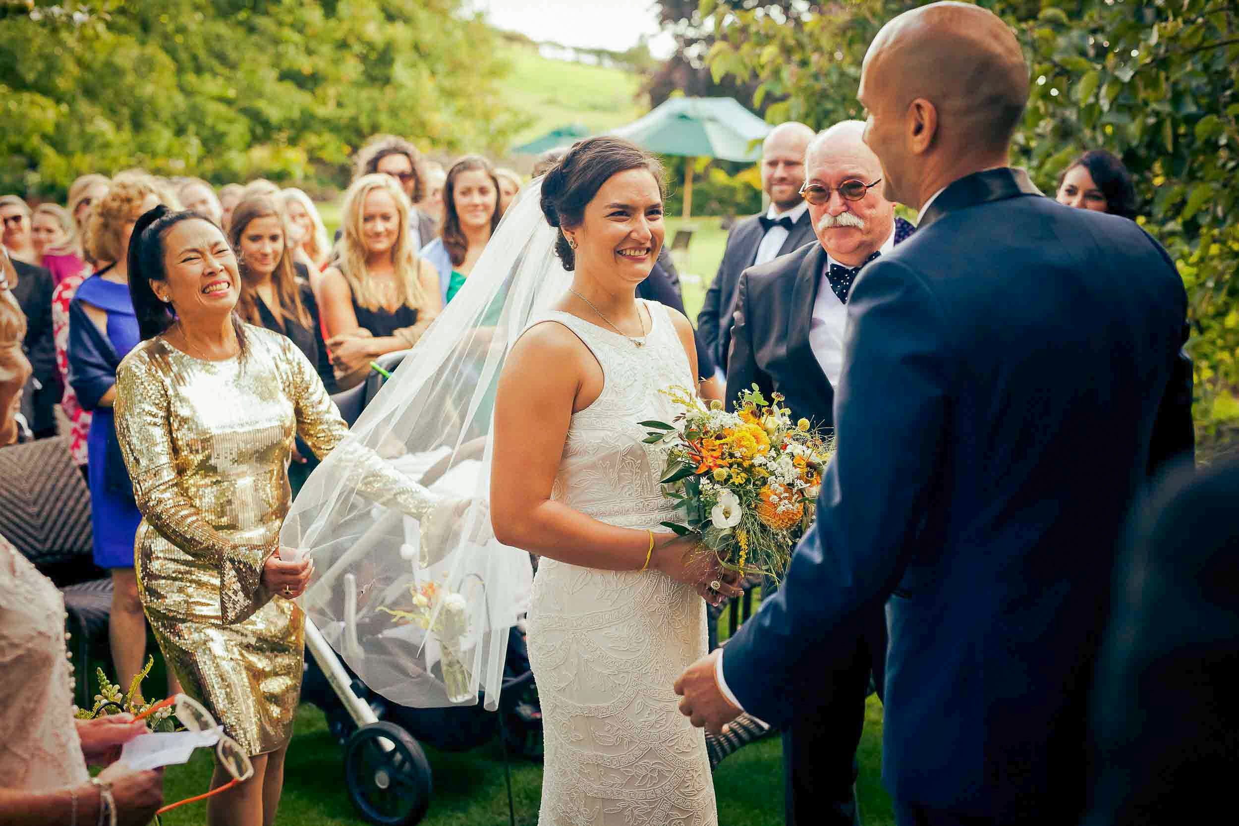 A bride and groom exchanging vows outdoors during a wedding ceremony, surrounded by guests and greenery.