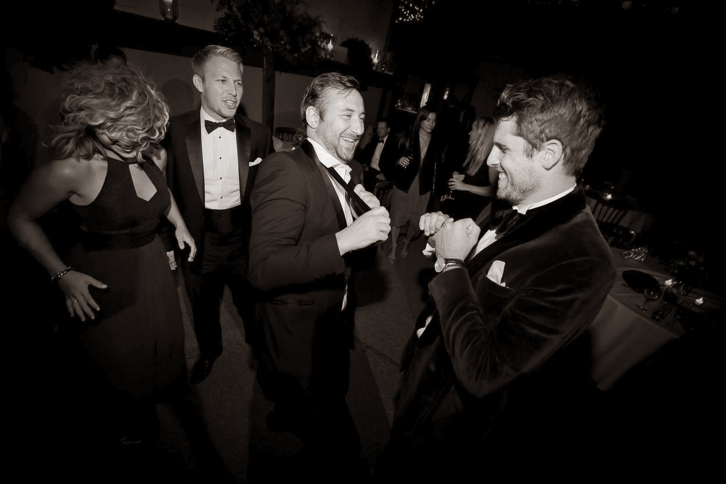 Two men in tuxedos, smiling and playing with their fists, face each other in a lively party setting.