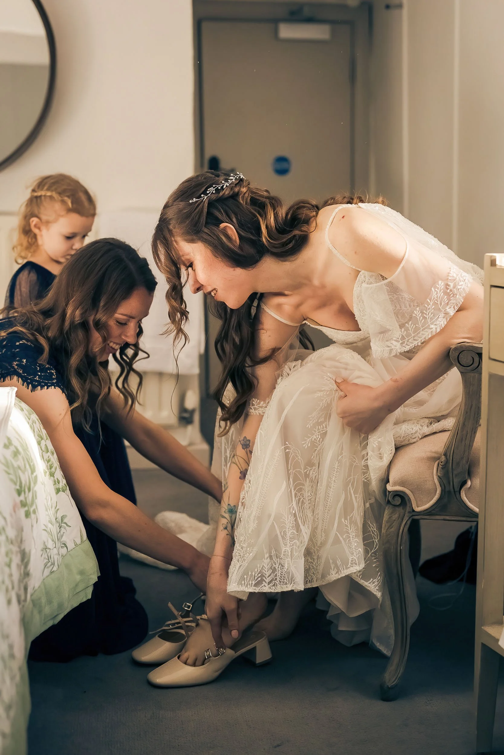 A bride in a white wedding dress sits on a chair while her bridesmaids help her put on beige high heels.