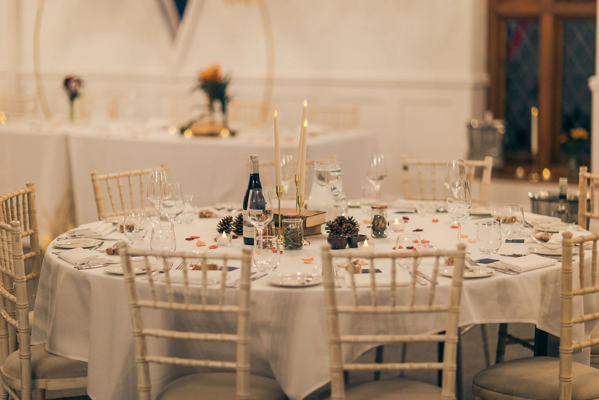 Round banquet table decorated with pinecones, candles, wine bottles, and scattered flower petals, set for a formal event with multiple glasses, plates, and silverware, surrounded by elegant chairs.