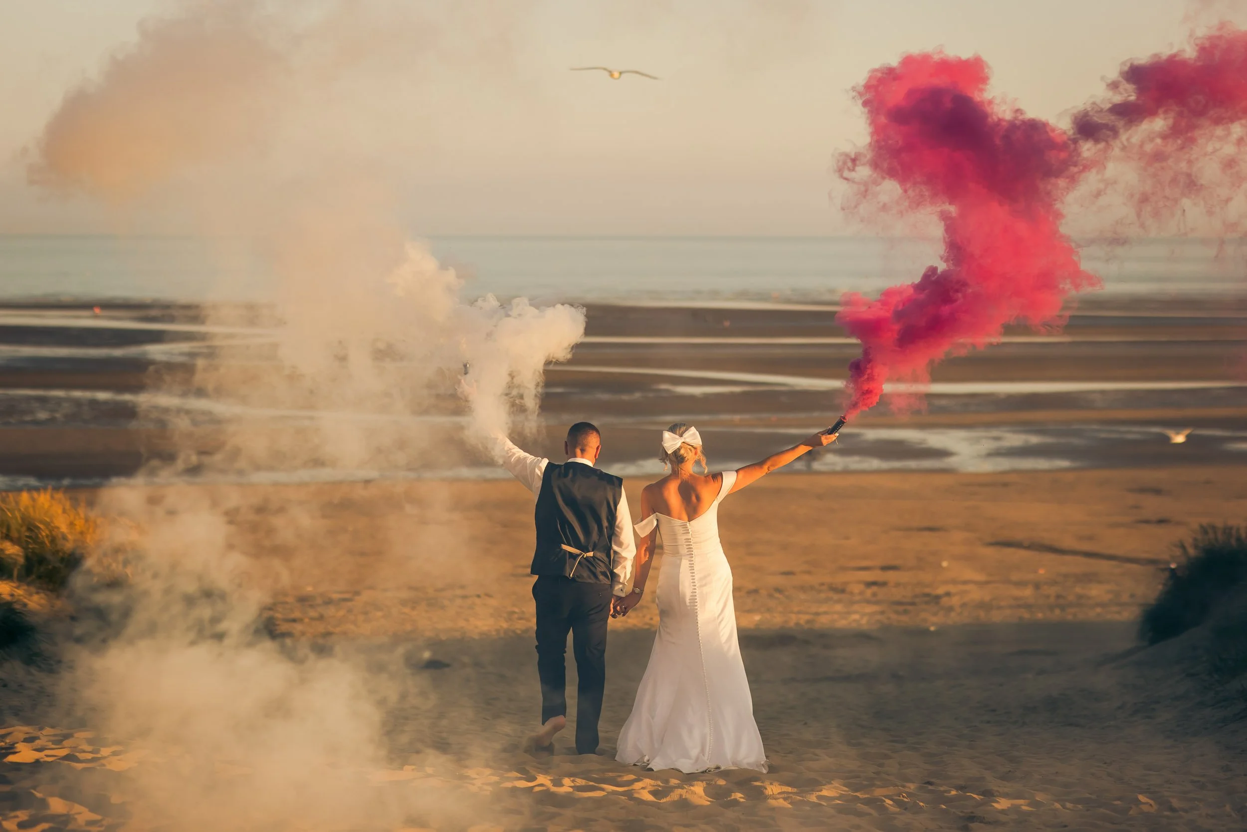 A couple in wedding attire holding hands on the beach, with the bride holding pink smoke and the groom holding white smoke, during sunset.