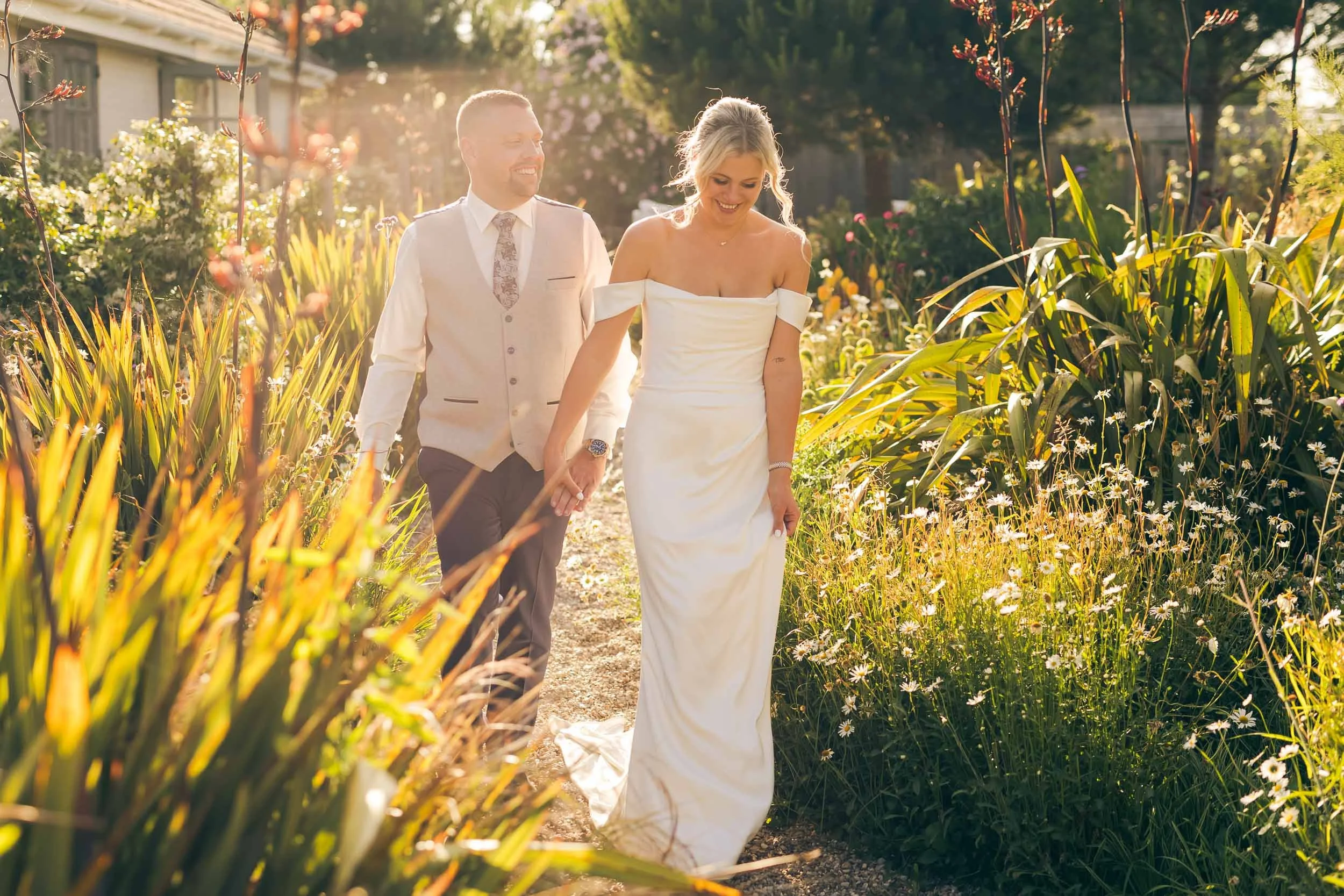 A bride and groom walking hand in hand on a garden path during sunset, surrounded by tall wildflowers and greenery.