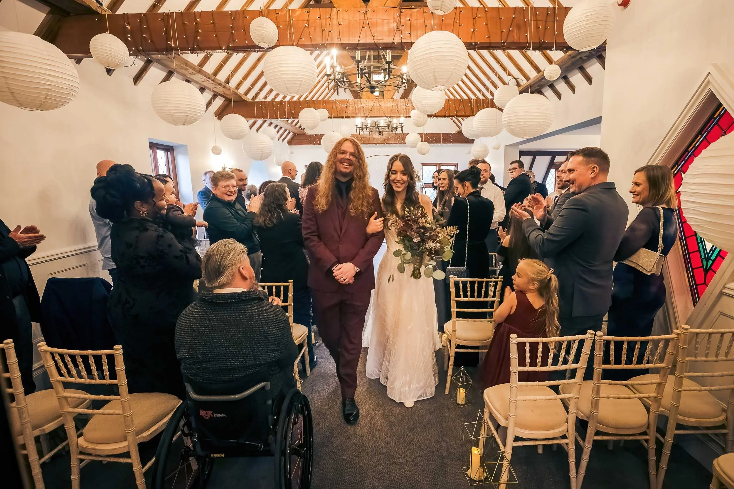 Wedding ceremony with a bride and groom walking down aisle while guests applaud indoors decorated with paper lanterns and string lights.