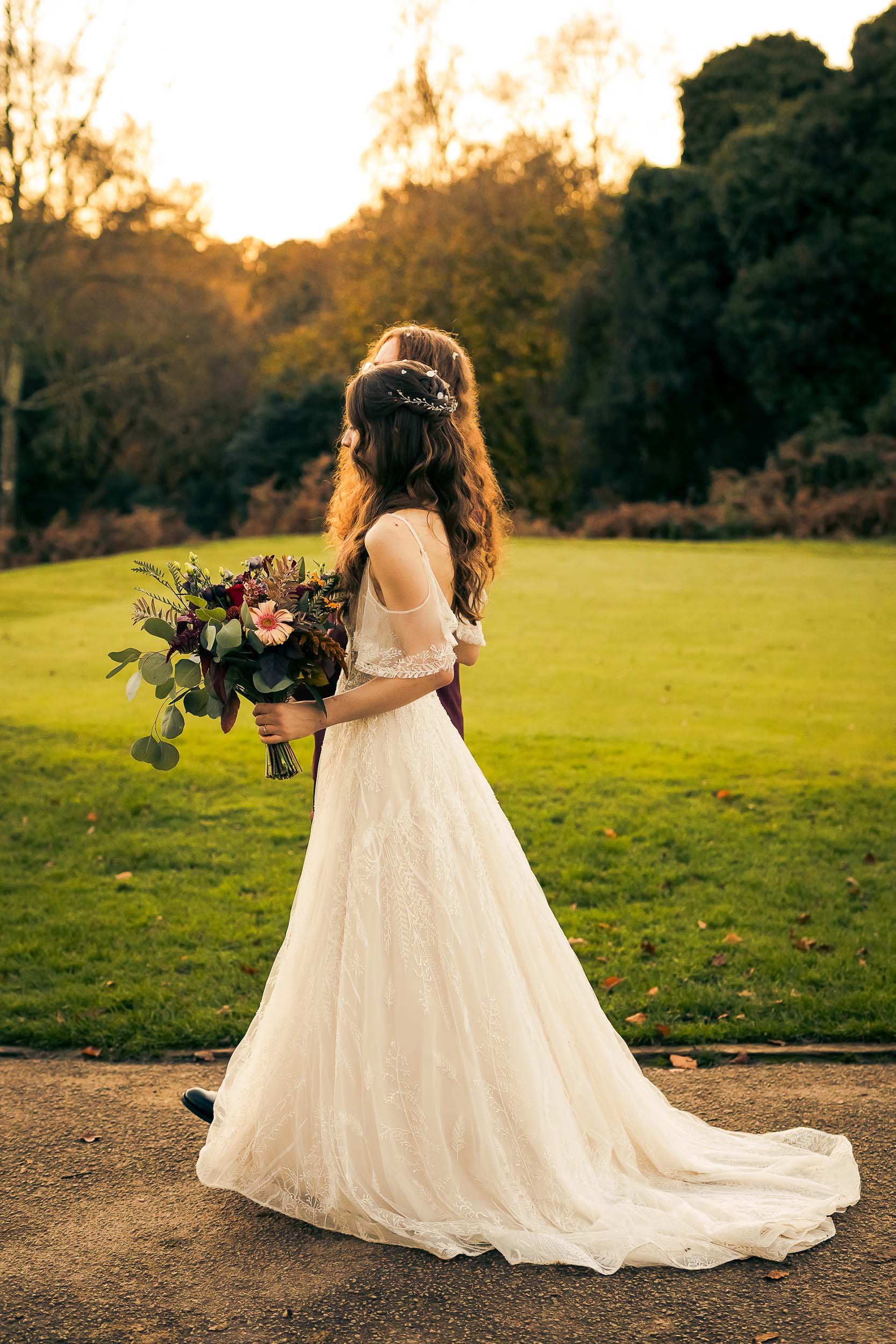 A bride holding a bouquet of flowers stands on a pathway outdoors at sunset, wearing a white lace wedding dress with off-the-shoulder sleeves and a floral crown in her hair.