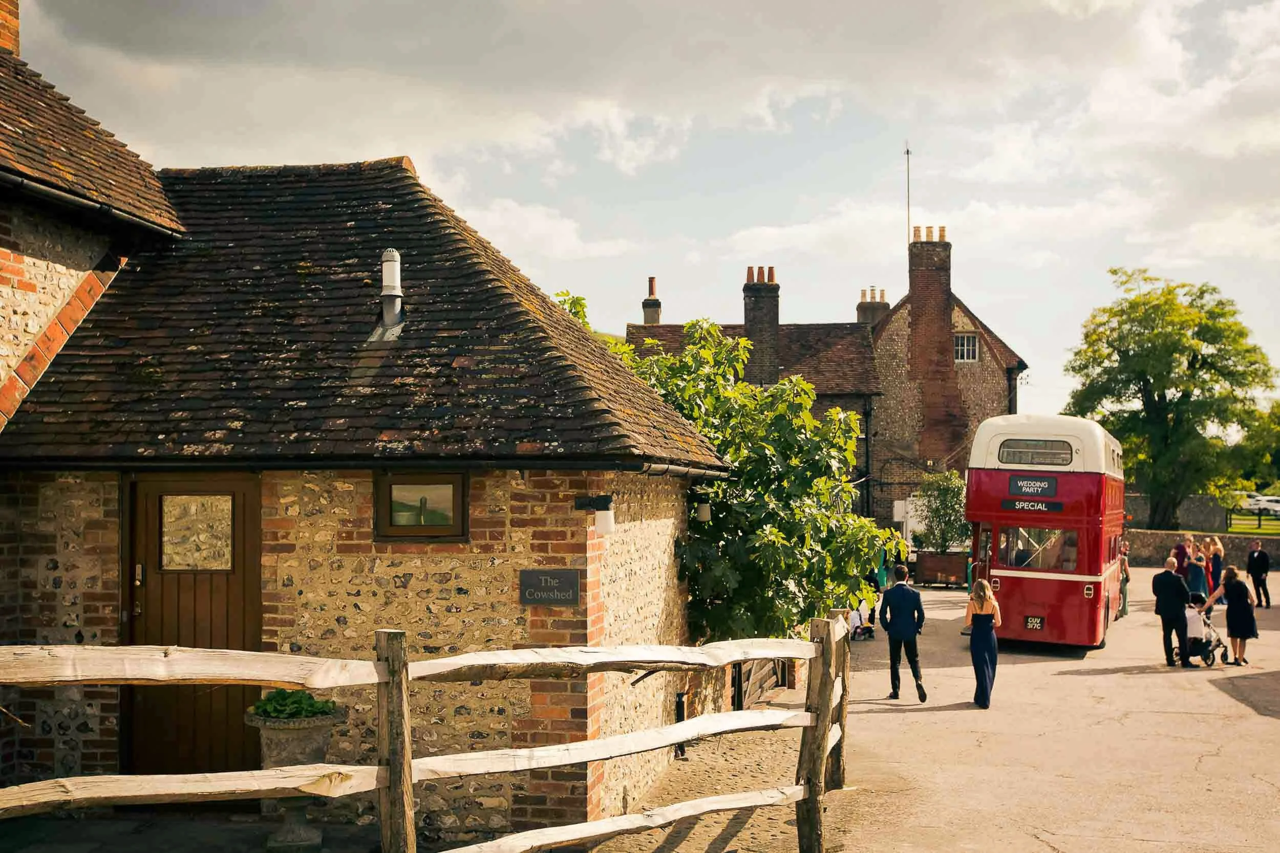 A historic brick building with a sign that reads 'The Cowshed' in a rural village, with a wooden fence in front, a red double-decker bus labeled 'Wedding Party Special,' and people walking around on a sunny day with trees and old buildings in the bac