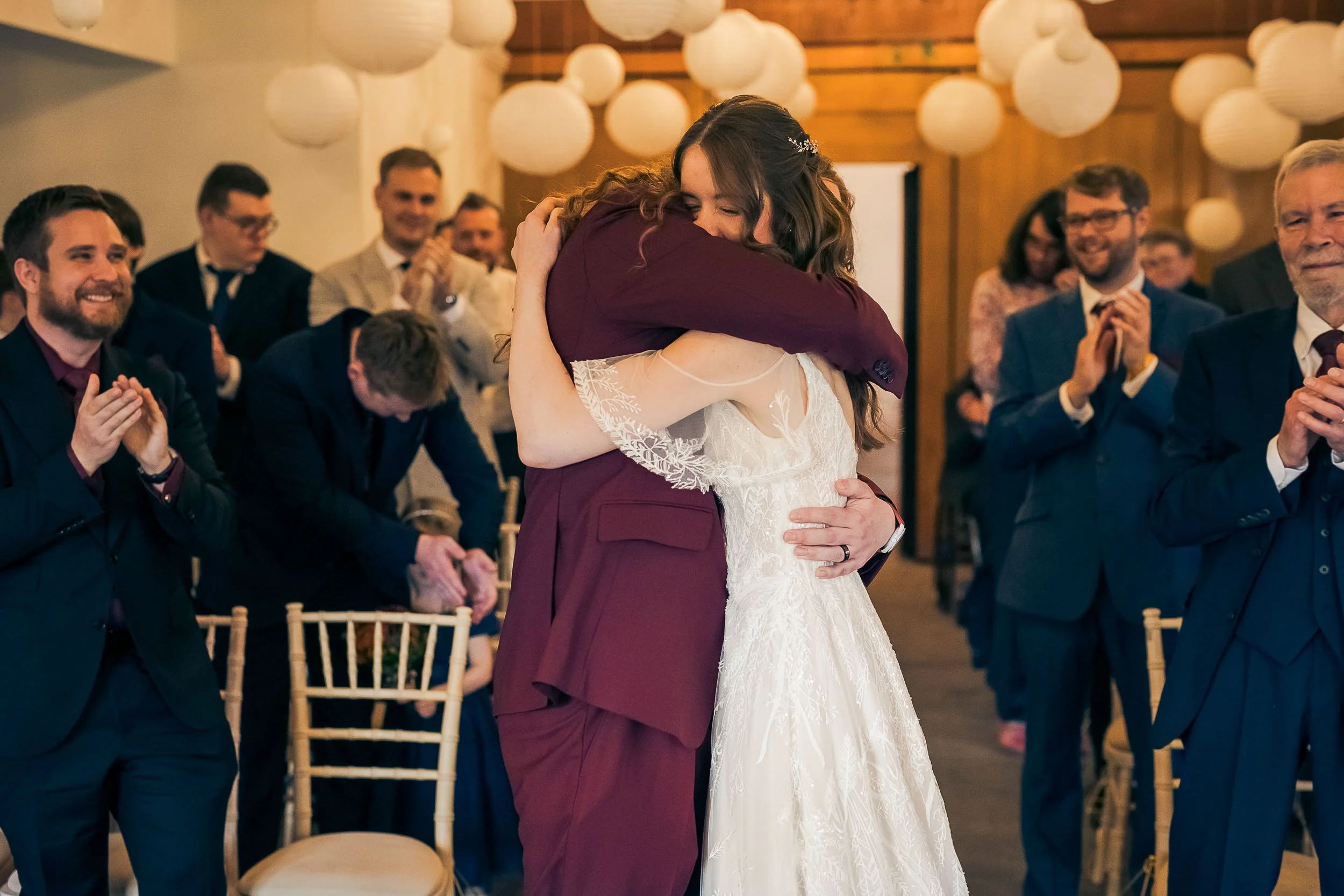A bride and groom embrace during their wedding ceremony, surrounded by seated and clapping guests in a decorated indoor venue.