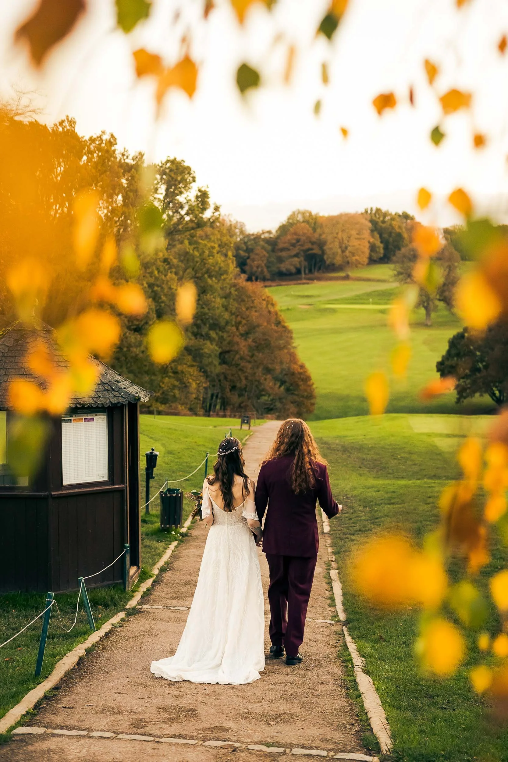 Two women, one in a wedding dress and the other in a purple suit, walk together on a path through a park surrounded by trees, during autumn.