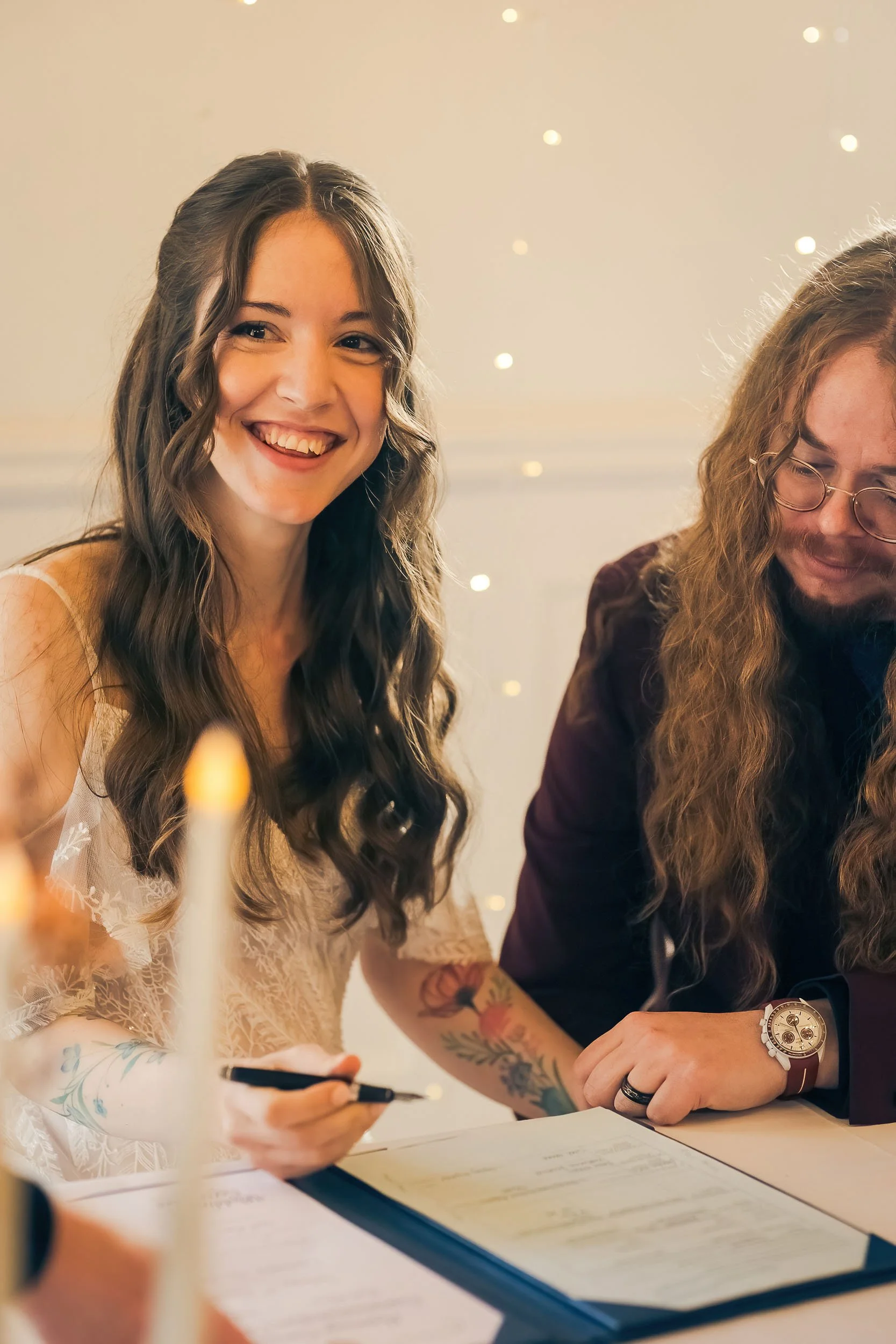 A young woman with long wavy hair smiling at a wedding signing a document, with a man with long curly hair and glasses next to her.