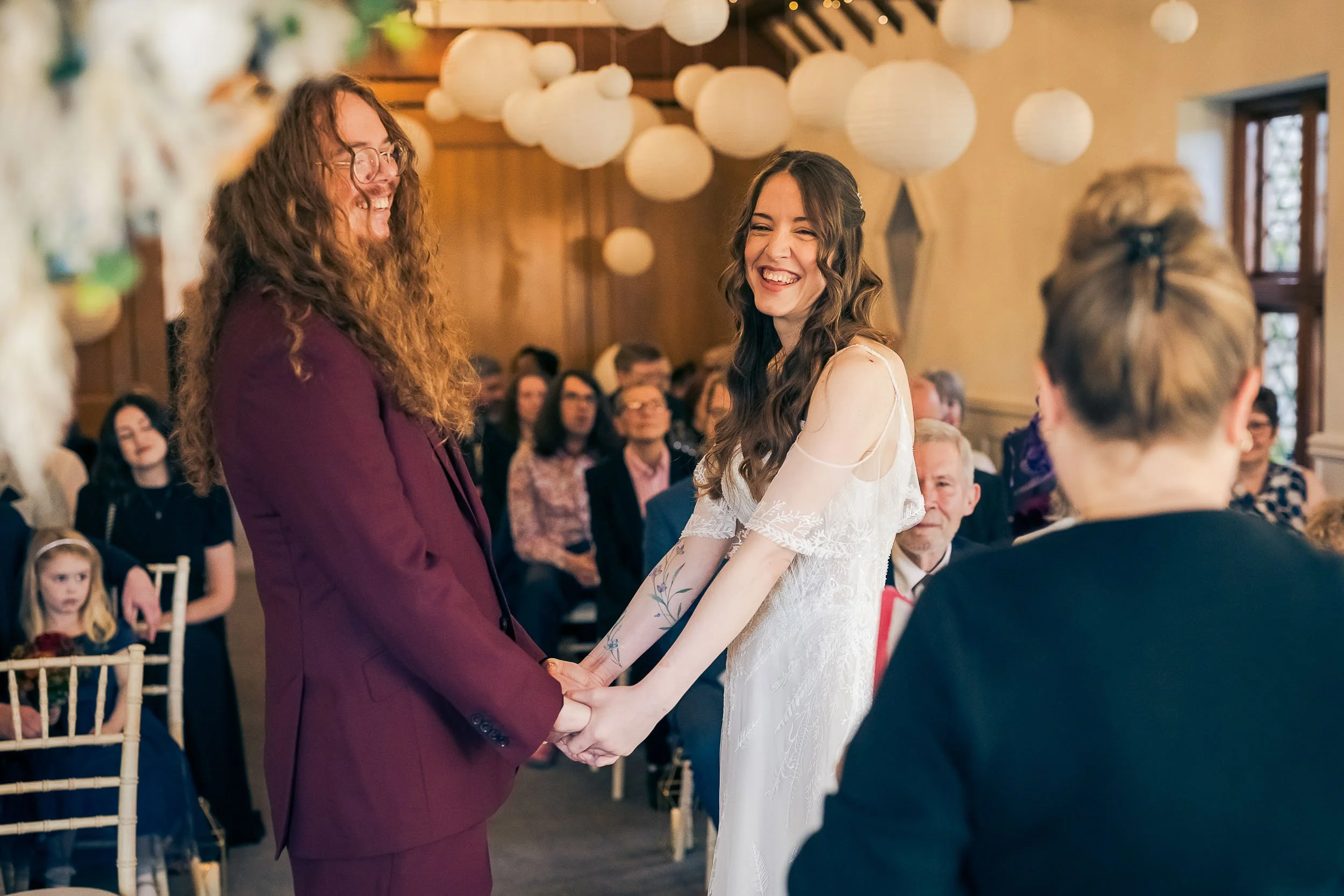 A smiling couple holding hands during a wedding ceremony in a decorated indoor venue filled with seated guests.
