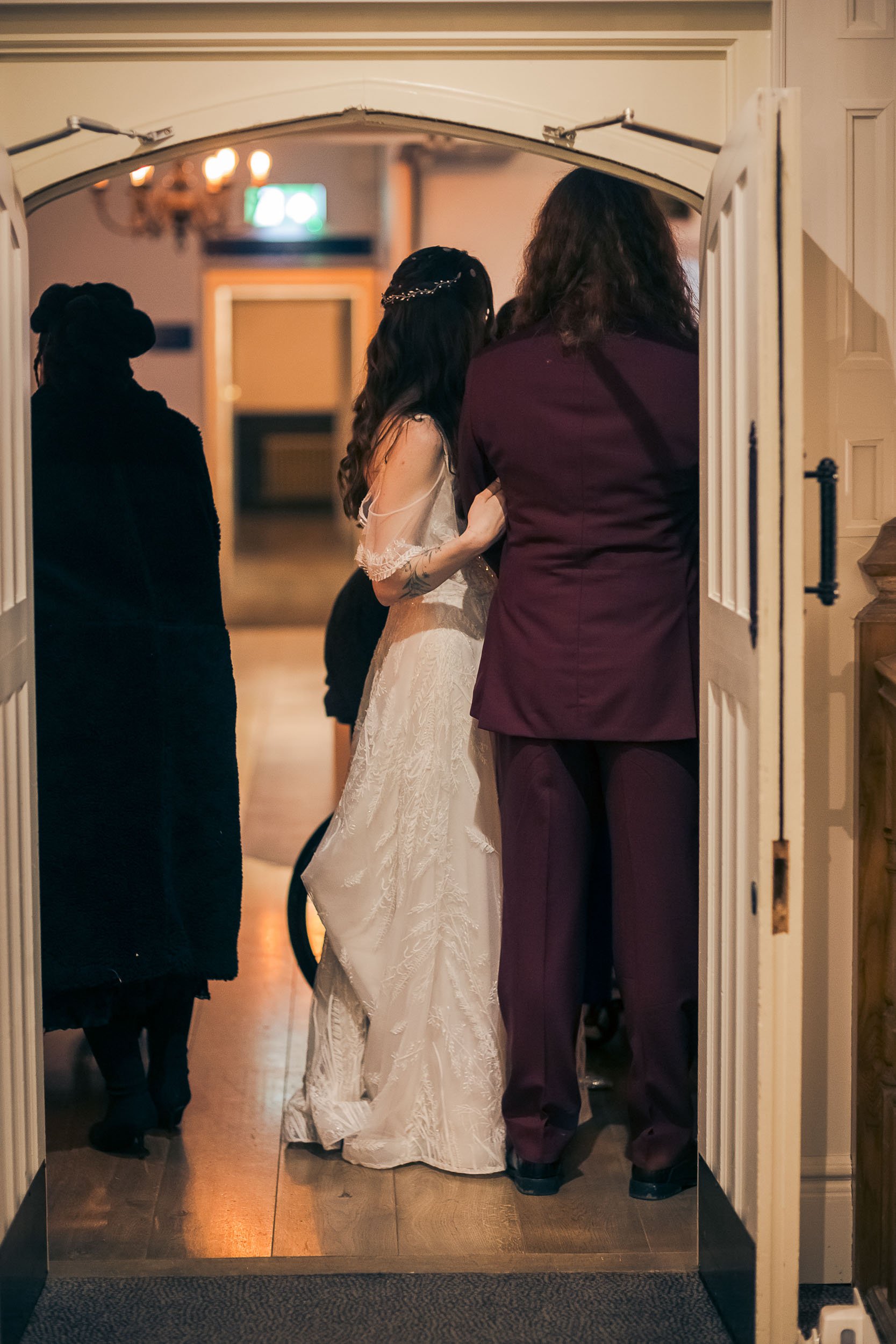 A bride in a white wedding dress walking into a room with a man in a maroon suit, with two women nearby, one in black. The scene appears to be at a wedding reception or ceremony.