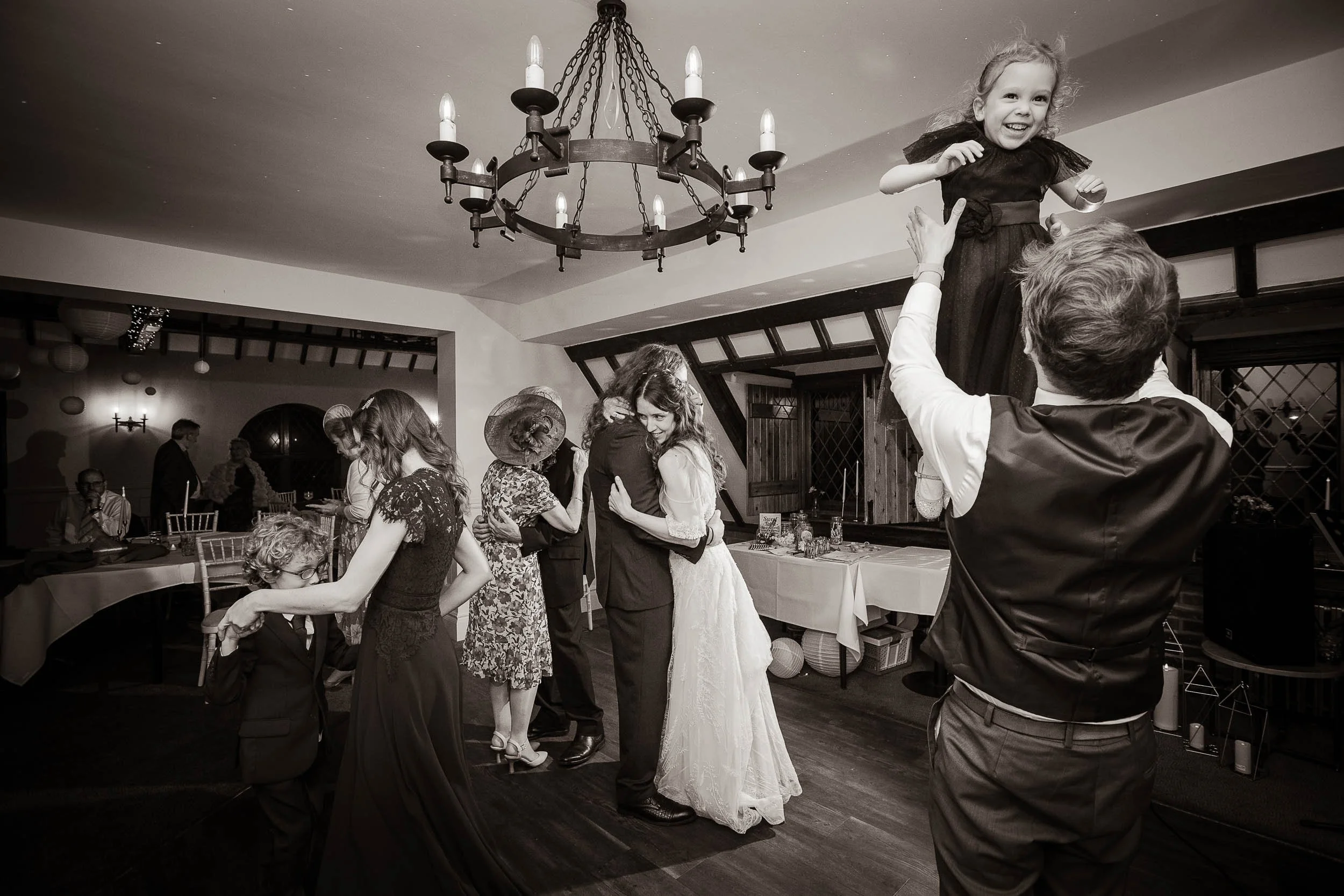 Black and white photo of a wedding reception with dancing guests, including a man lifting a young girl in a dress, a woman and man hugging, and a young boy holding a woman's hand, under a chandelier in a decorated indoor venue.