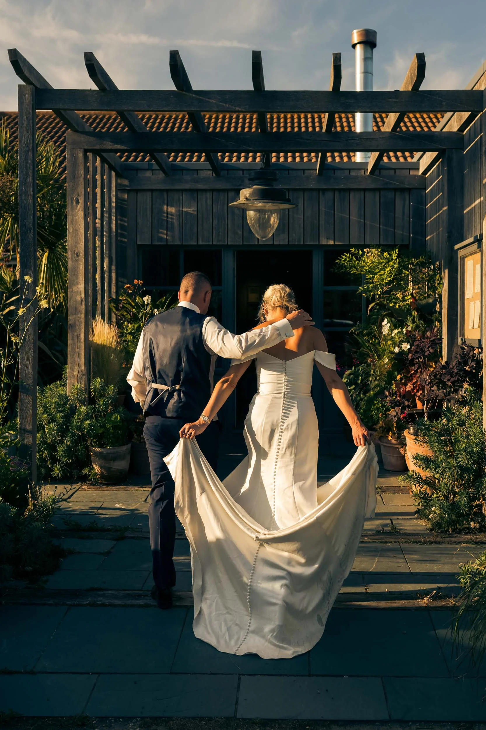A bride and groom walking outside on their wedding day at sunset, with the bride holding her dress and the groom gently touching her shoulder.